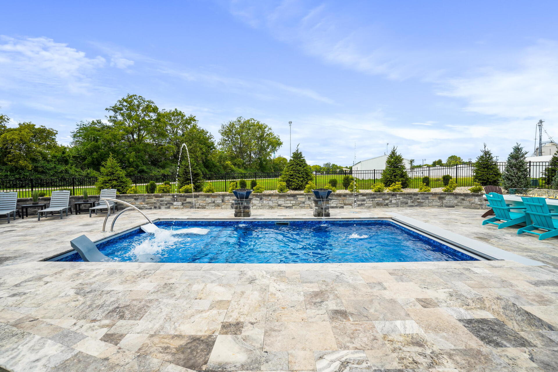 Outdoor swimming pool with blue water, stone deck, lounge chairs, and greenery under a bright sky
