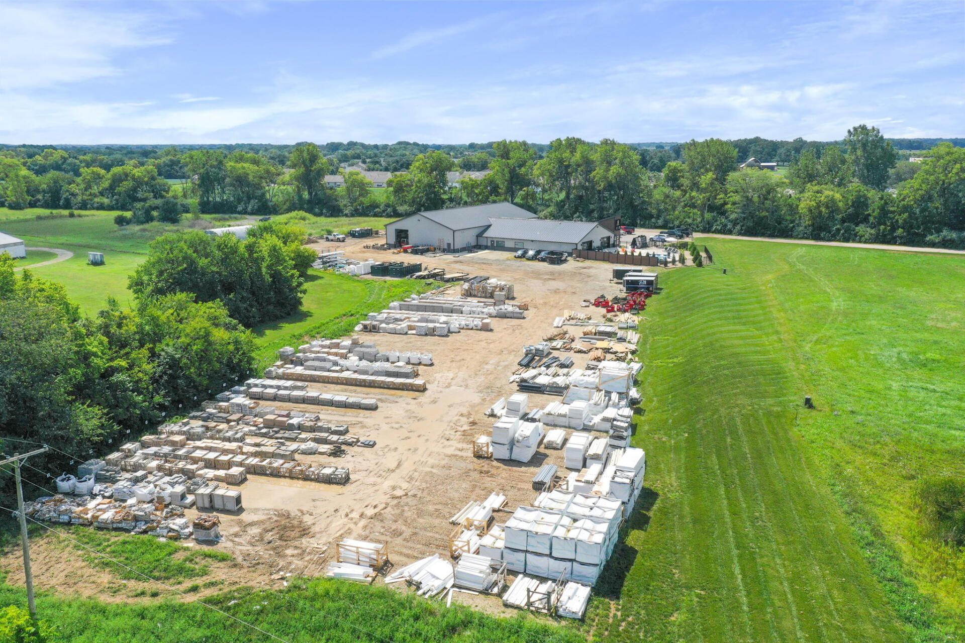 Aerial view of a lumber yard with stacks of wood, white materials, and a warehouse beside green fields.