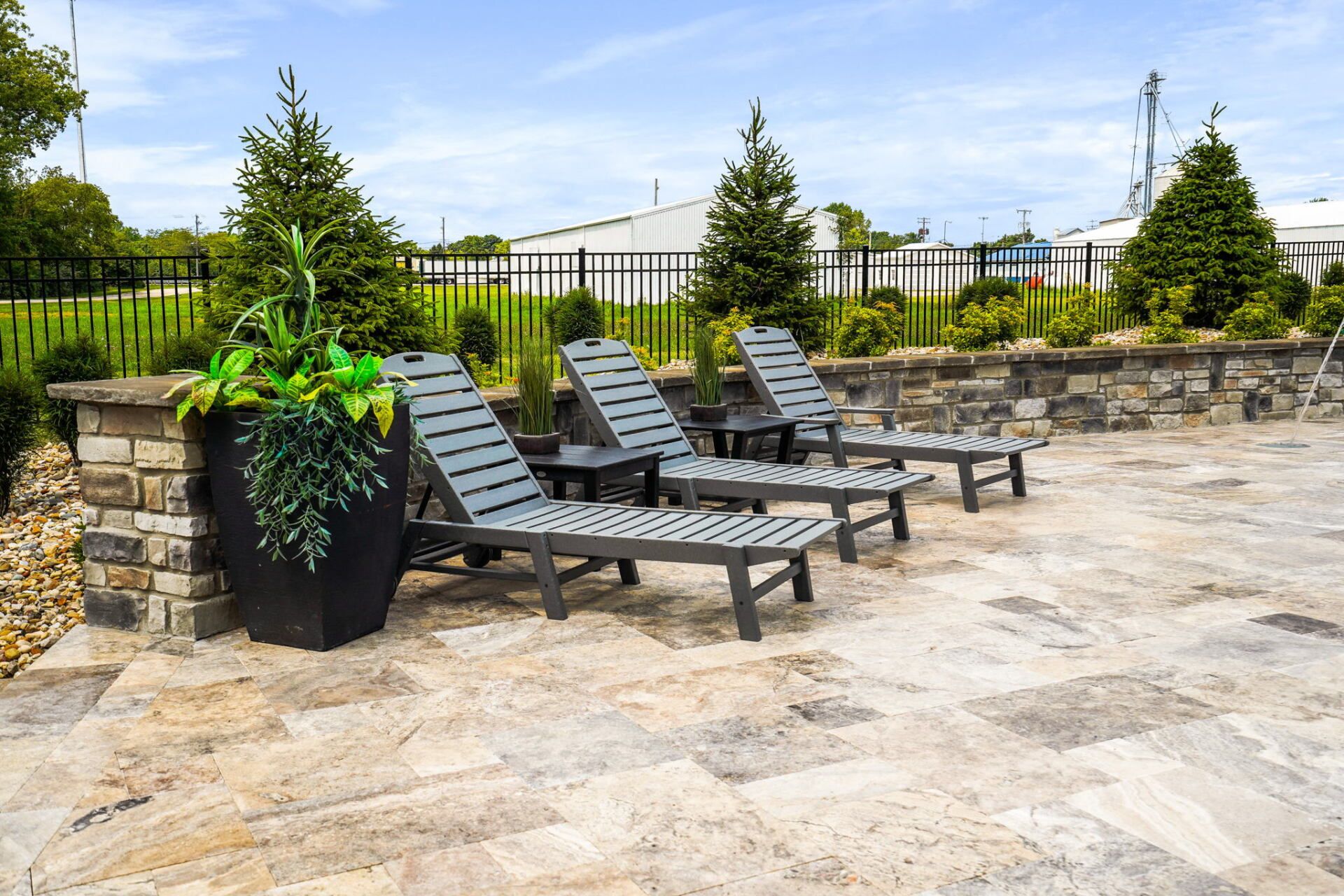 Stone patio with black lounge chairs, planters, and a stone wall in a landscaped outdoor area.