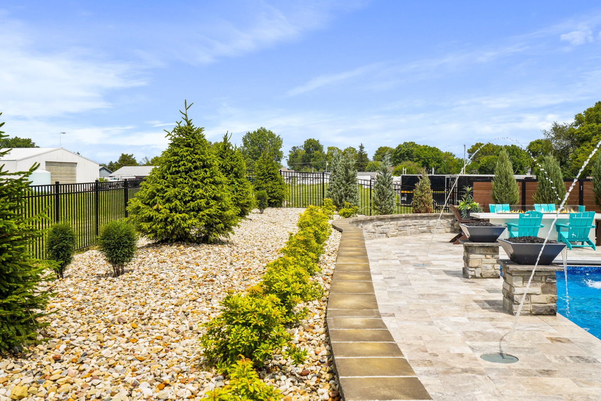 Landscaped backyard patio with stone walkway, shrubs, and turquoise chairs under a blue sky