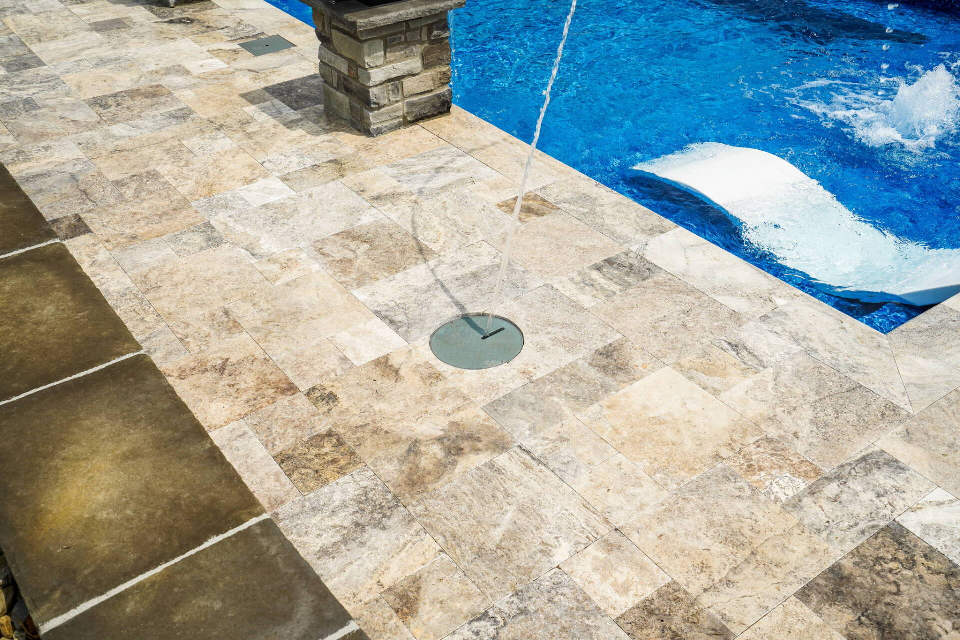 Stone pool deck beside a blue swimming pool, with a drain and a stone pillar.