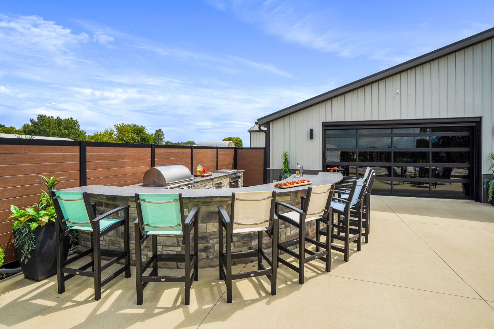 Outdoor patio with a long table, black chairs, potted plants, and a brick wall beside a modern building.