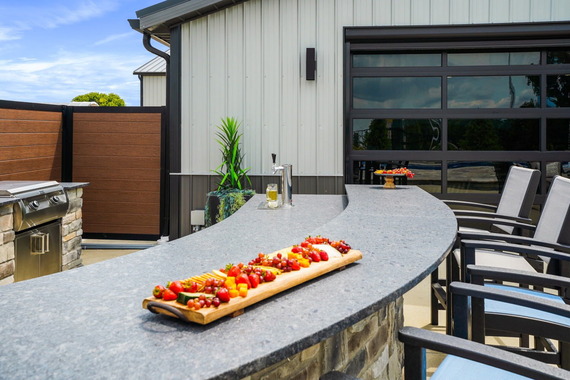 Modern backyard patio with curved stone bar, grill, black chairs, and a tray of food