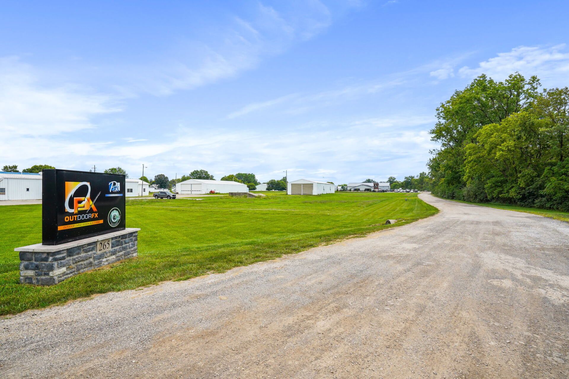 Gravel road curving past a grassy lot and trees under a blue sky, with a sign at the entrance