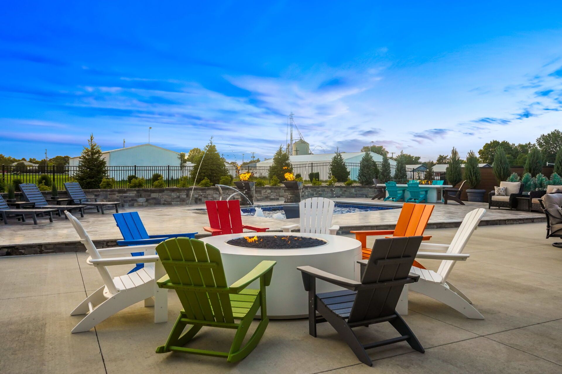 Colorful Adirondack chairs around a fire pit on a sunny outdoor patio, with trees and buildings in the background