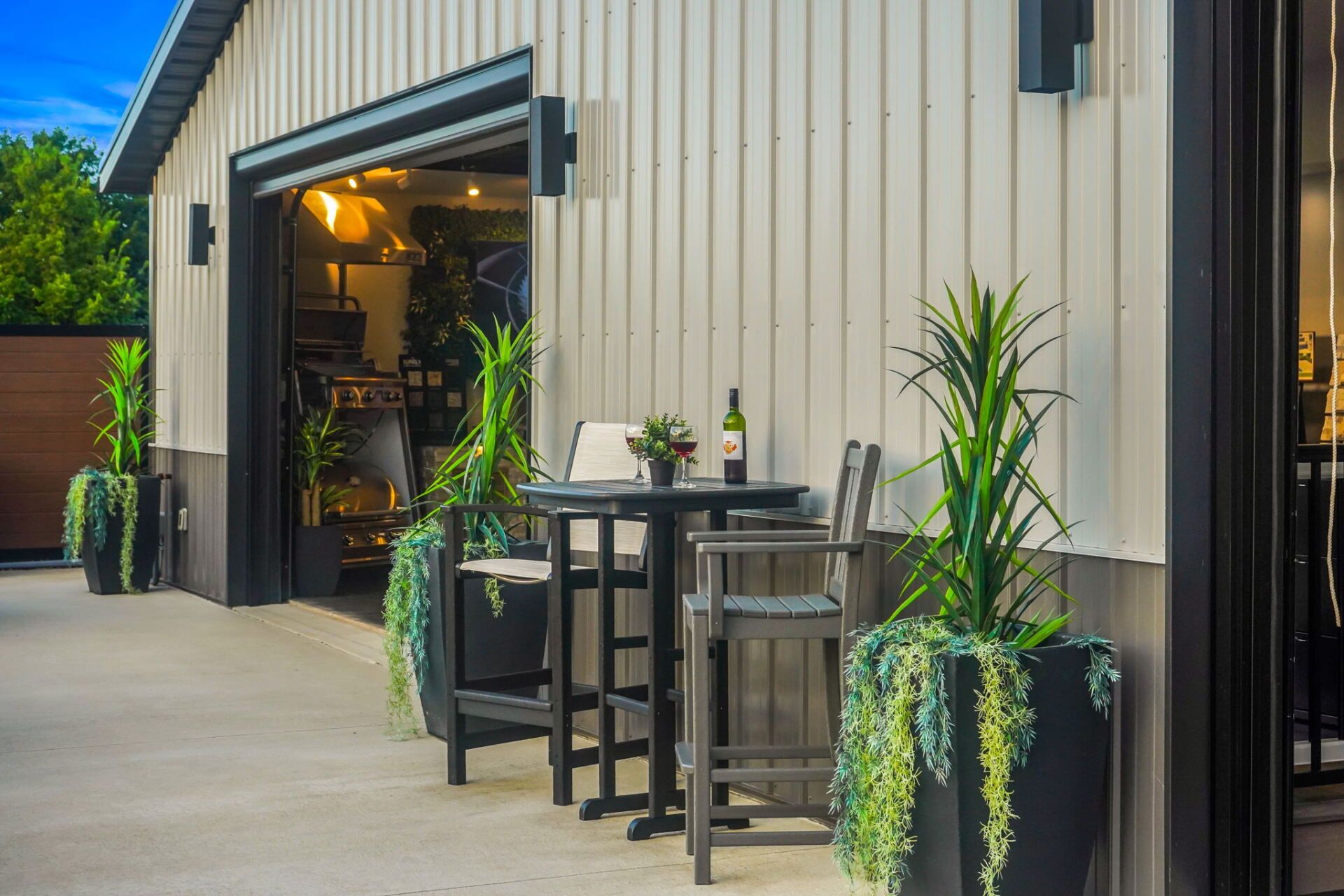 Outdoor café seating beside a beige corrugated building with potted plants and open garage-style doors