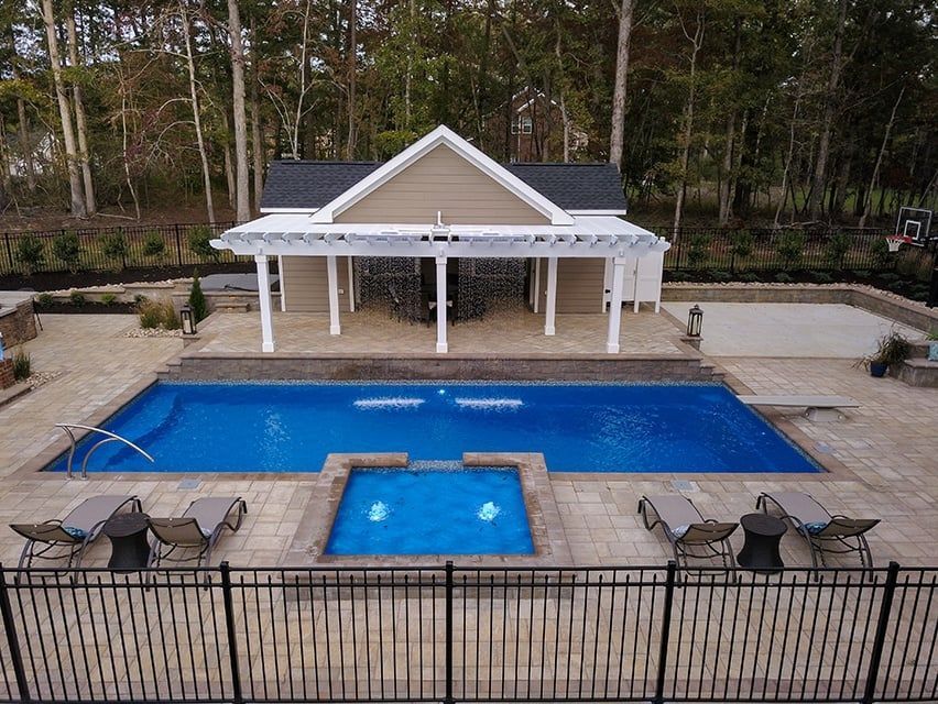 Backyard pool with blue water, hot tub, patio chairs, and a covered pavilion surrounded by trees