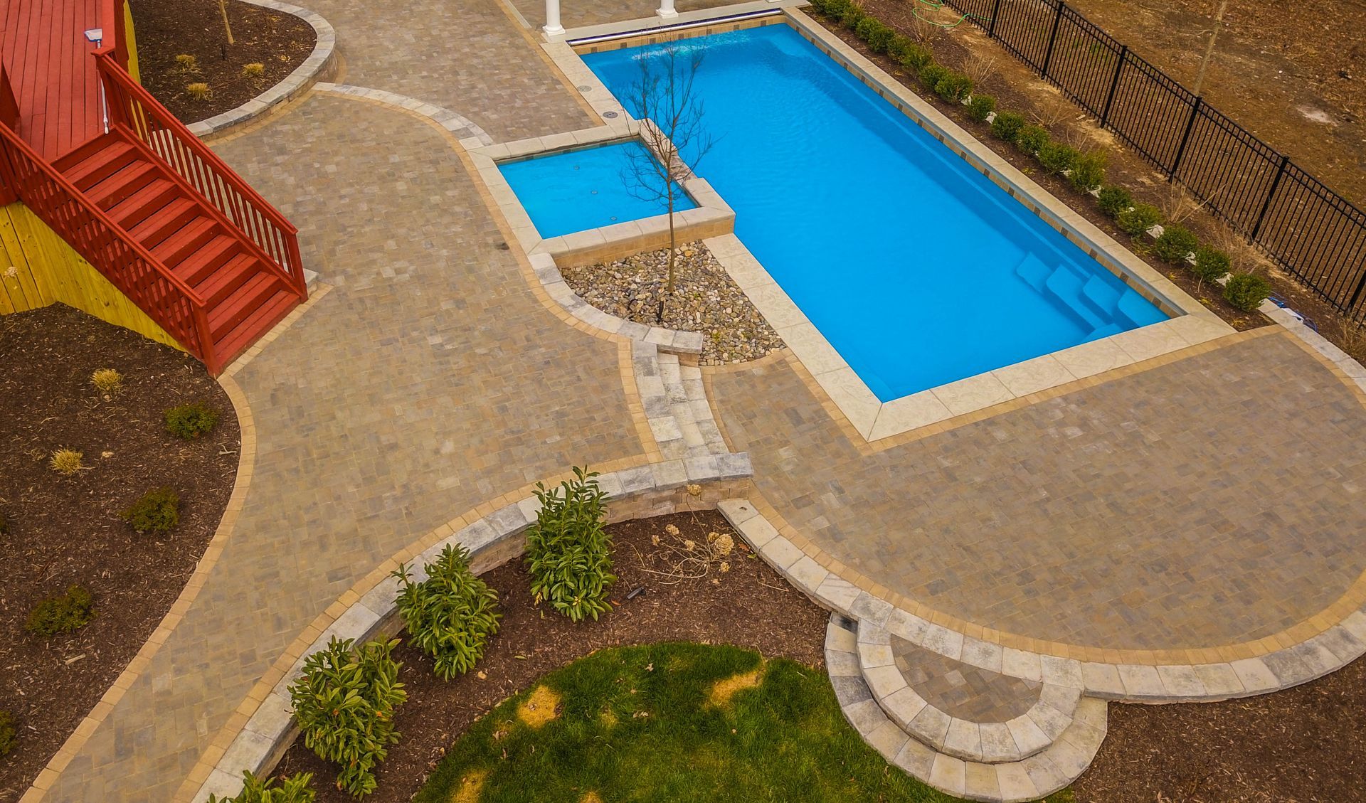 Aerial view of a patio with a bright blue pool, stone walkways, and red stairs beside landscaped greenery