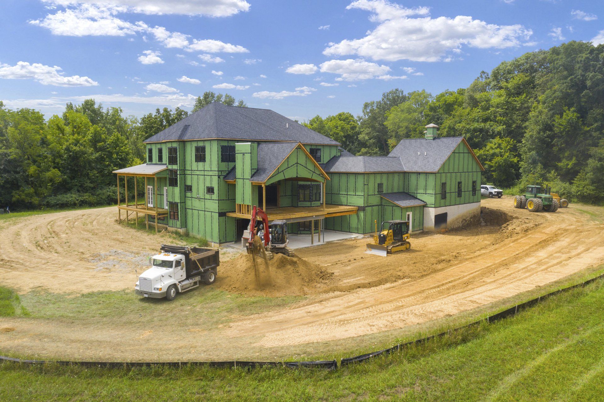 Green house under construction on a dirt lot, with workers, trucks, and trees under a blue sky