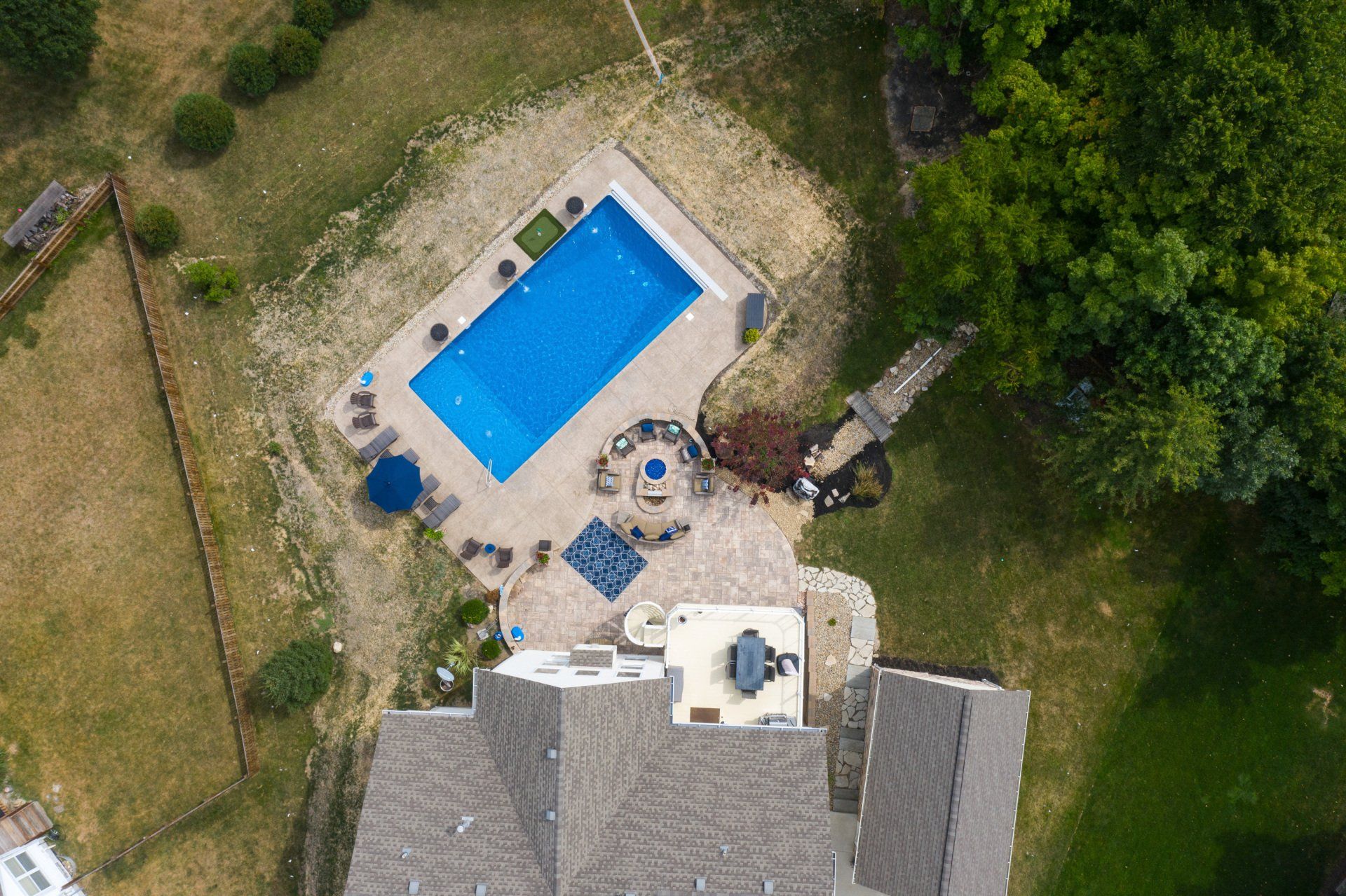 Aerial view of a backyard with a blue rectangular pool, patio furniture, and a house beside grassy trees.