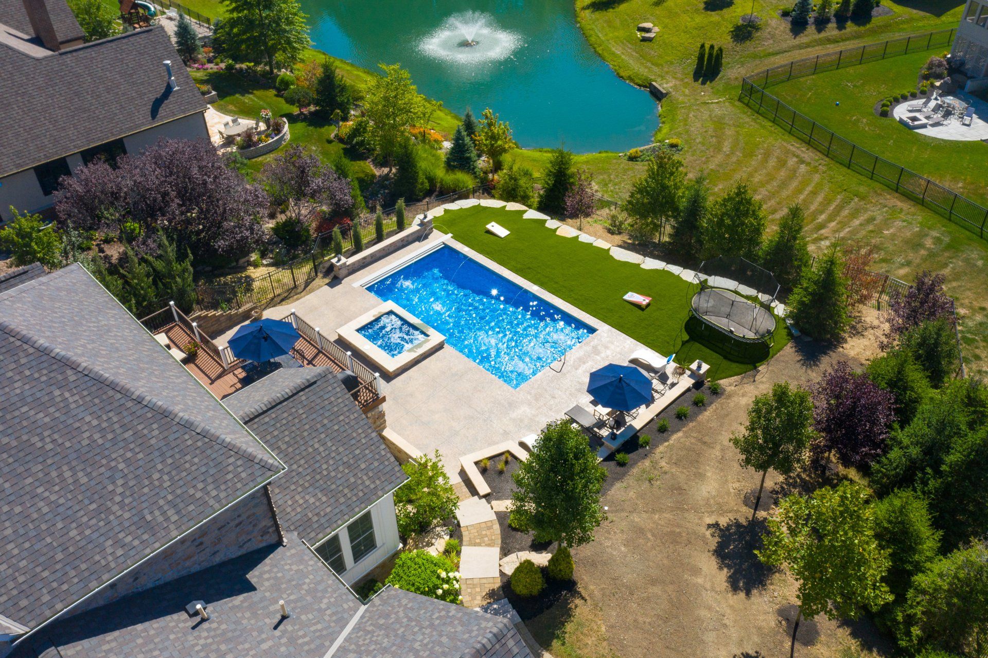 Aerial view of a backyard pool, hot tub, patio, and surrounding lawn with a pond nearby.