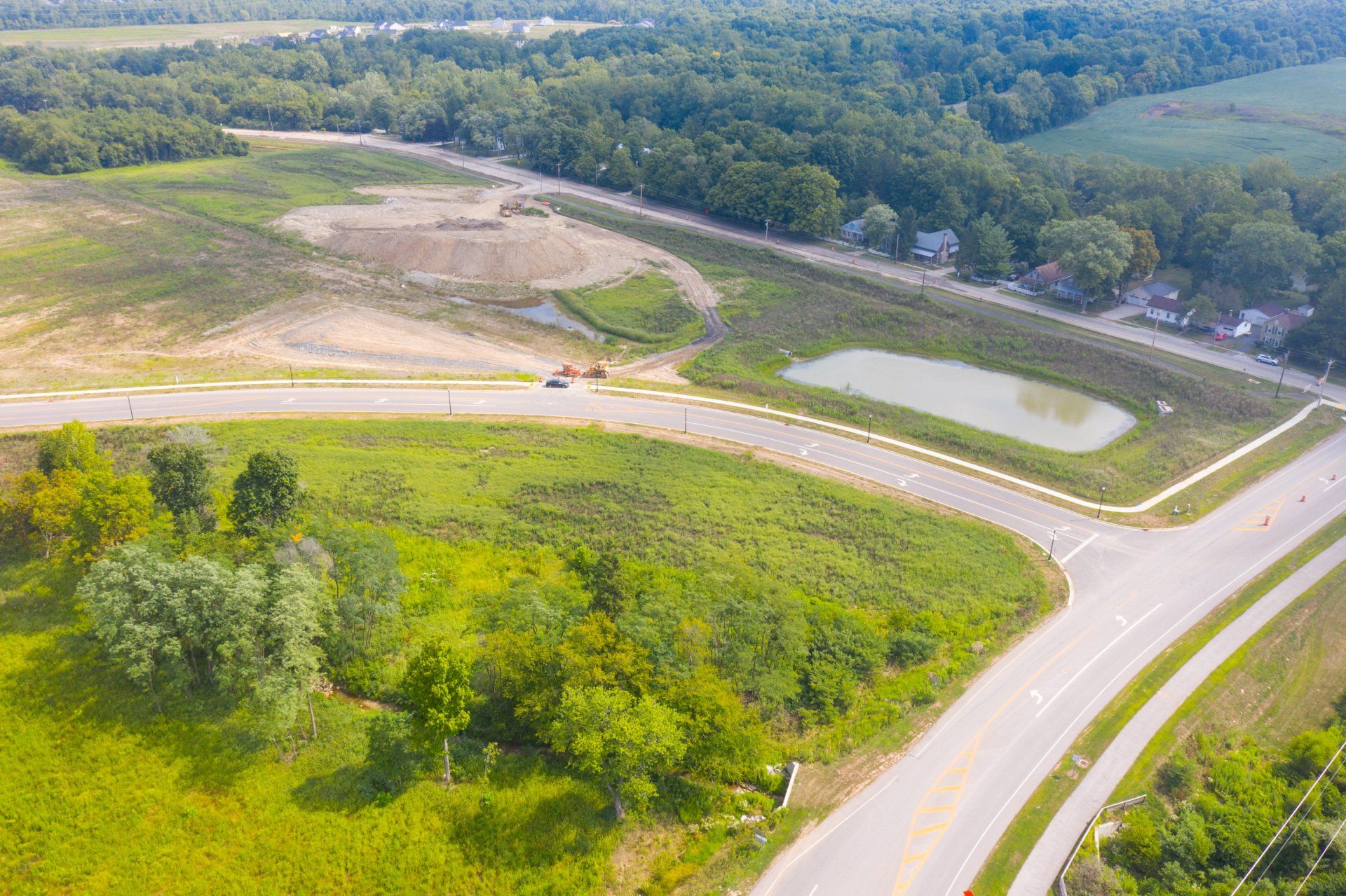 Aerial view of a rural road curving past a small pond, open fields, and dense green trees