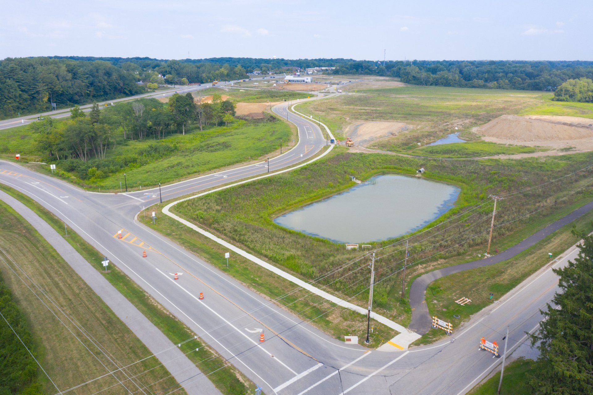 Aerial view of a vacant road junction with a pond, grassy lots, and winding roads under a blue sky