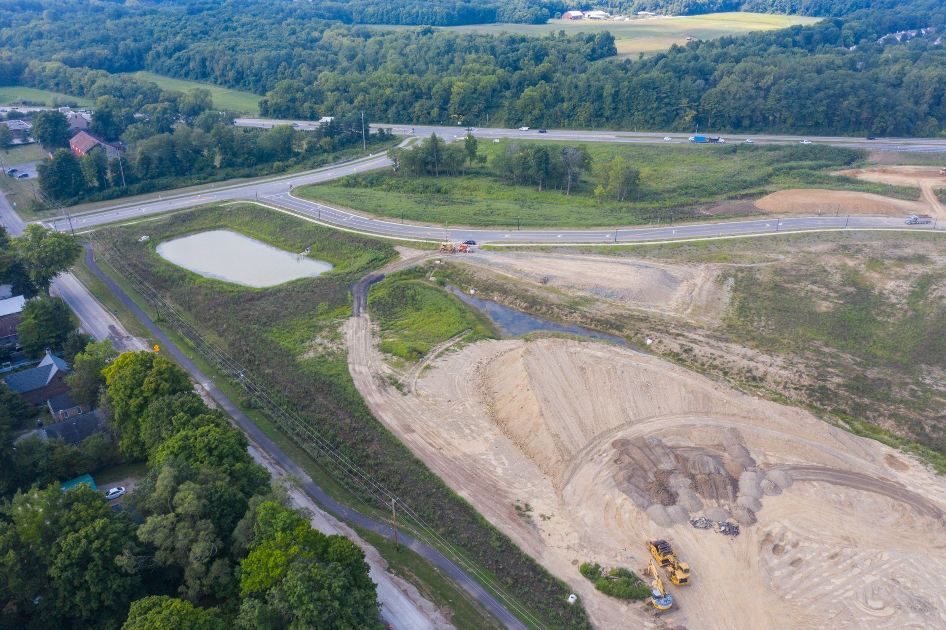 Aerial view of a construction site beside a road and trees, with dirt piles and a small pond