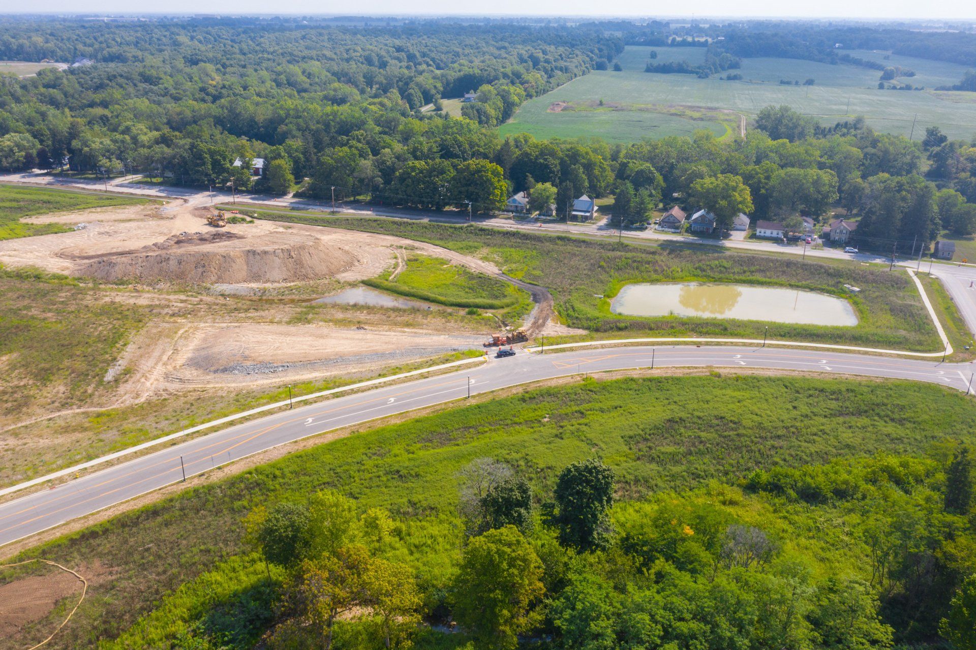 Aerial view of a road curving past grassy fields and a wooded area under clear daylight