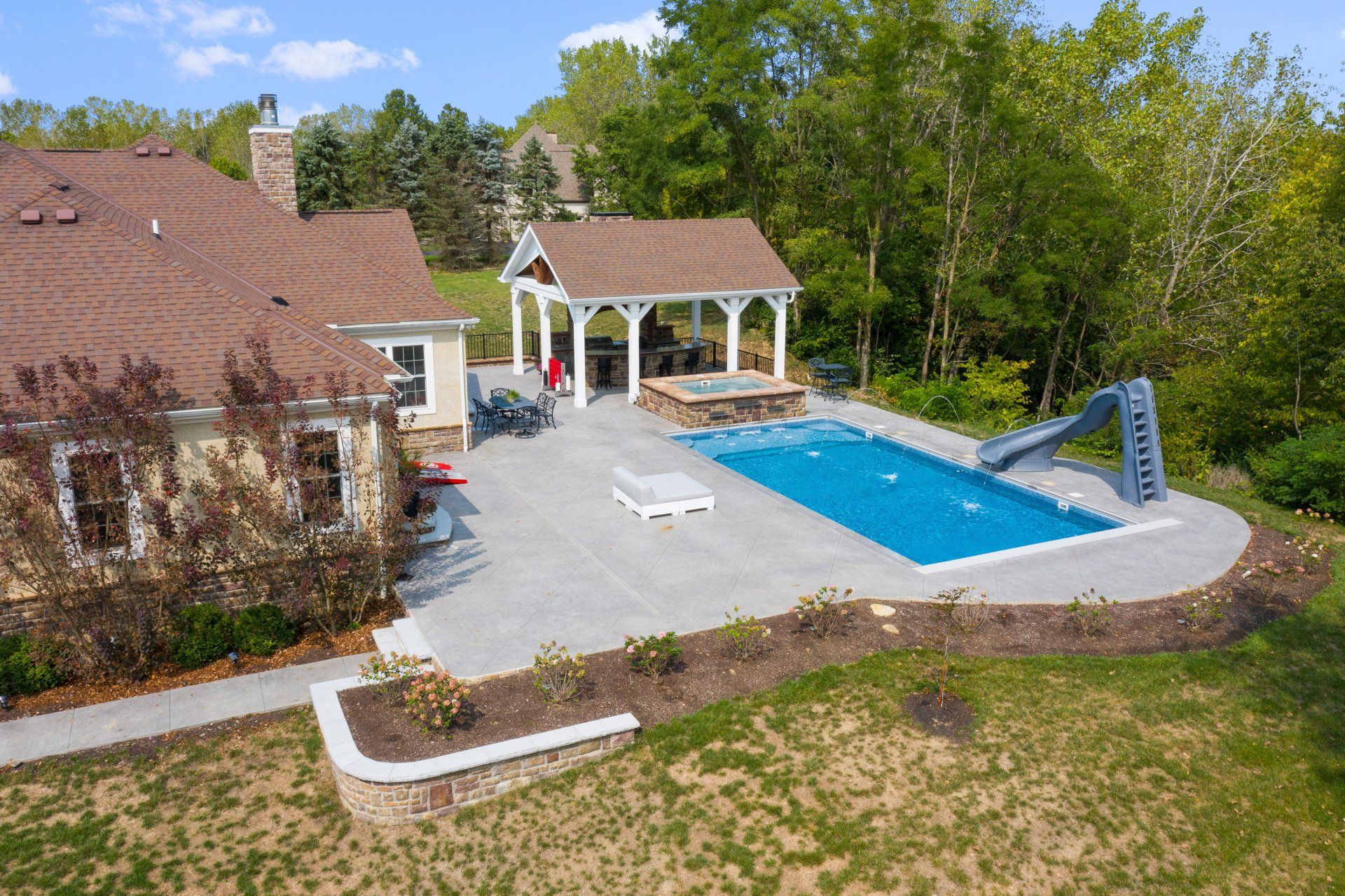 Backyard with a blue swimming pool, stone patio, pergola, and surrounding trees