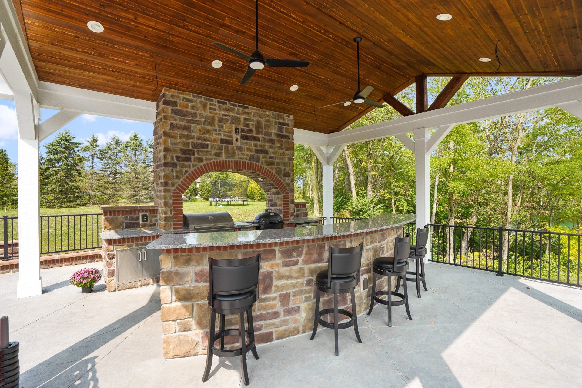 Covered outdoor bar with stone counter, barstools, and wood ceiling on a sunny patio