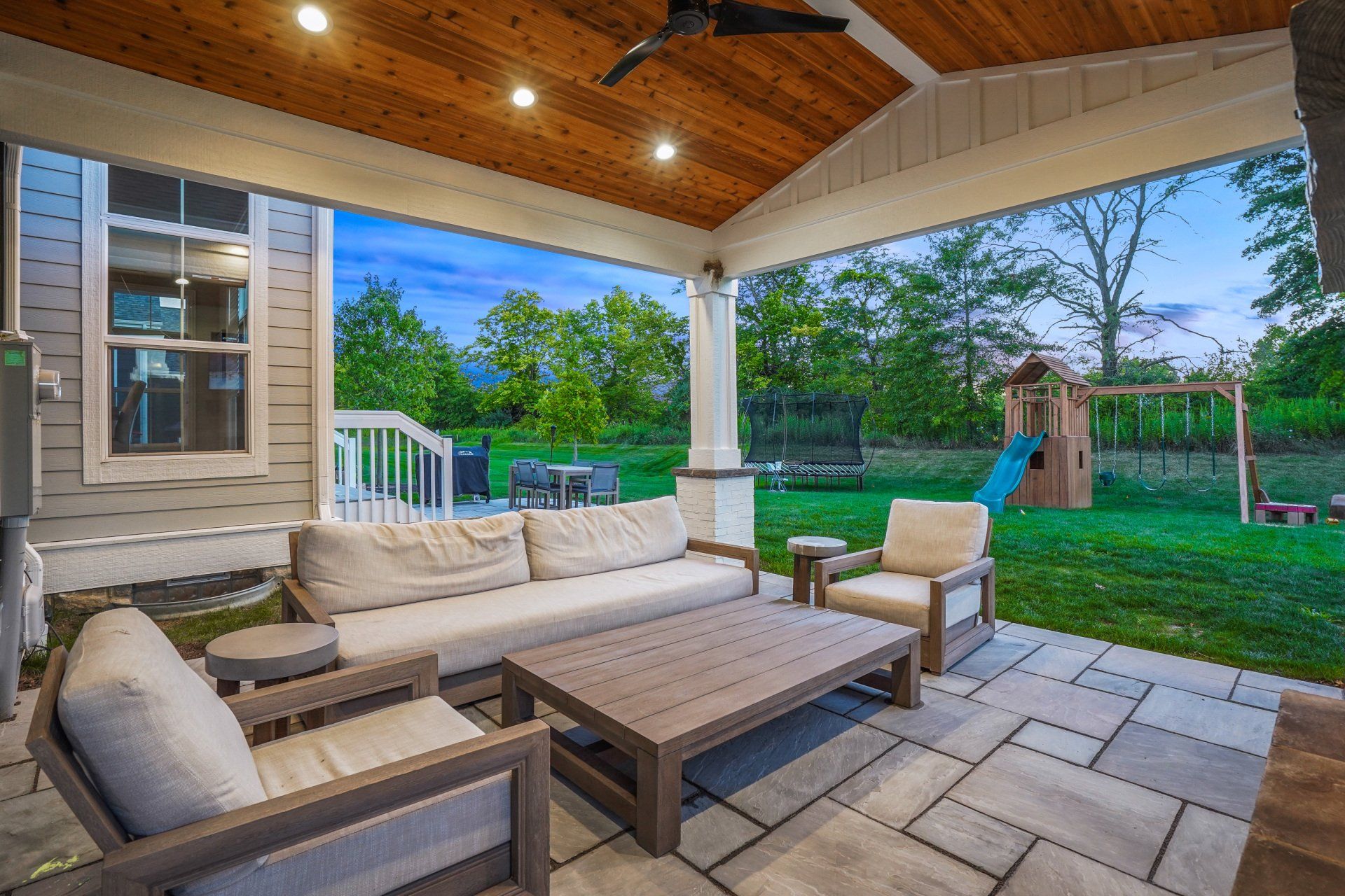Covered patio with wicker seating, wood ceiling, and a view of a green backyard and playset.