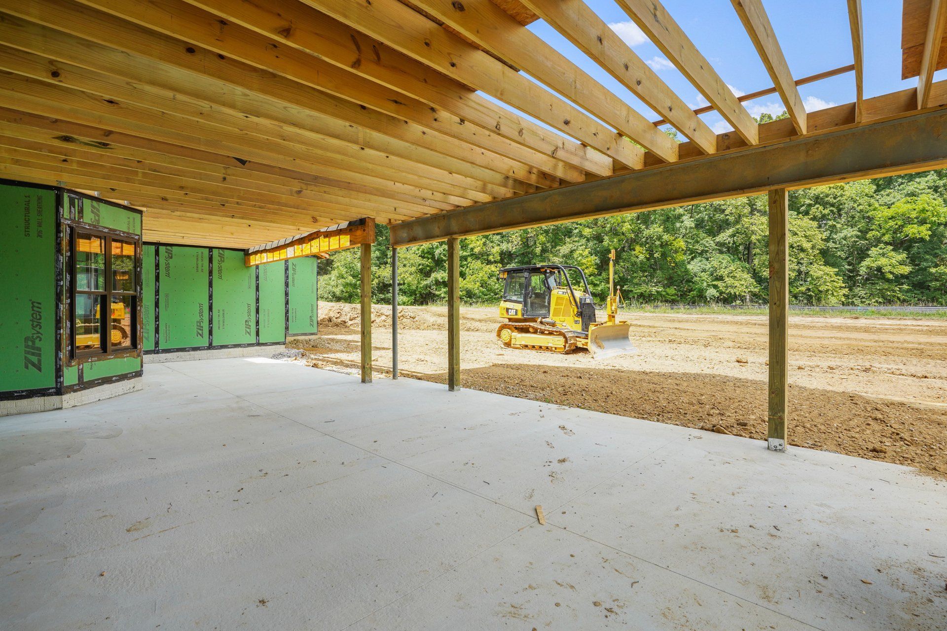 Partially built house with exposed beams, green sheathing, and a bulldozer on a dirt lot.