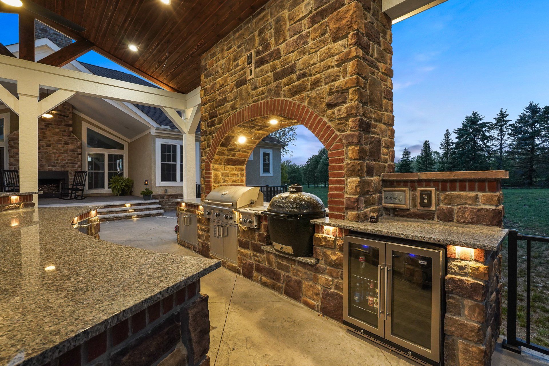 Stone outdoor kitchen with granite countertop, brick oven, and built-in grill on a covered patio at dusk