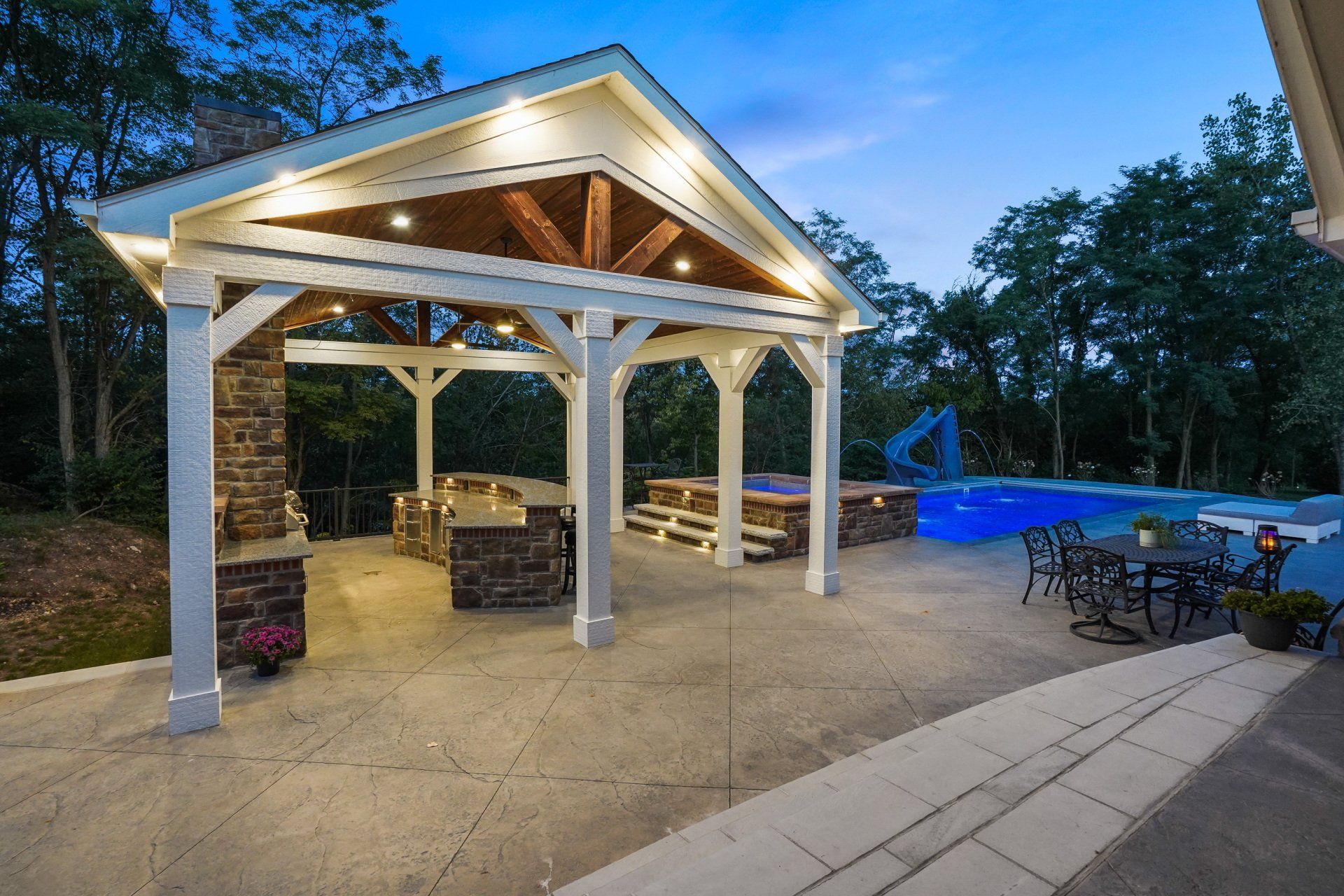 Covered outdoor pavilion with stone columns, lit ceiling, seating, and a poolside patio at dusk