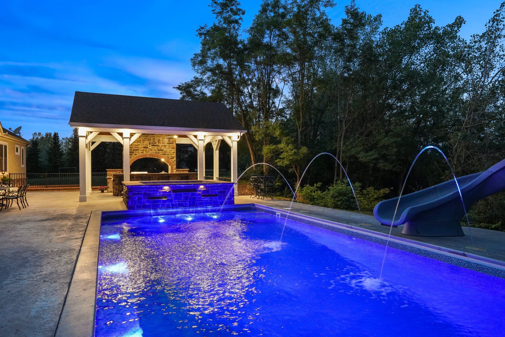 Illuminated backyard pool at dusk with blue water, gazebo, and slide