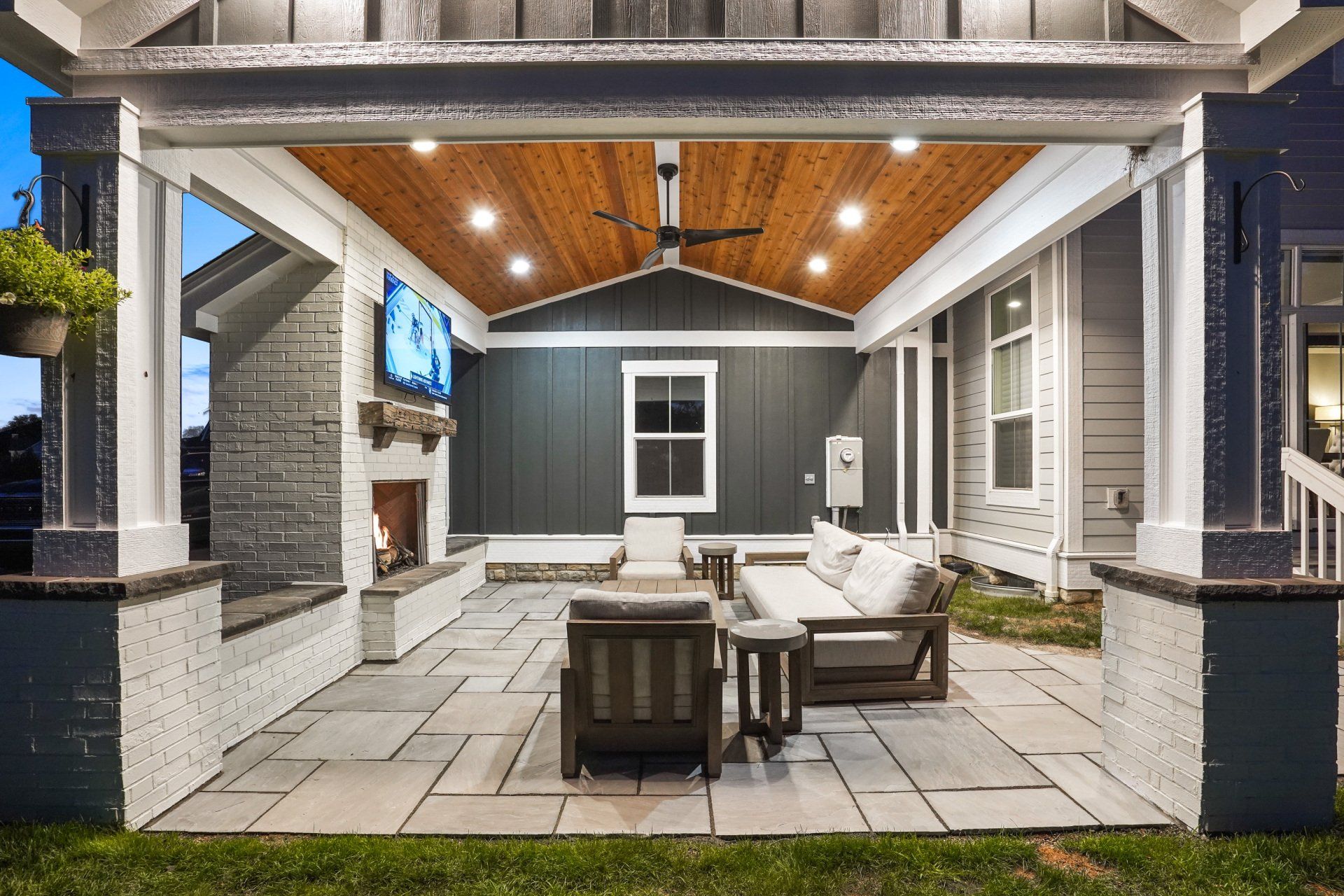 Covered backyard patio with stone pavers, white outdoor seating, and a wooden ceiling at dusk