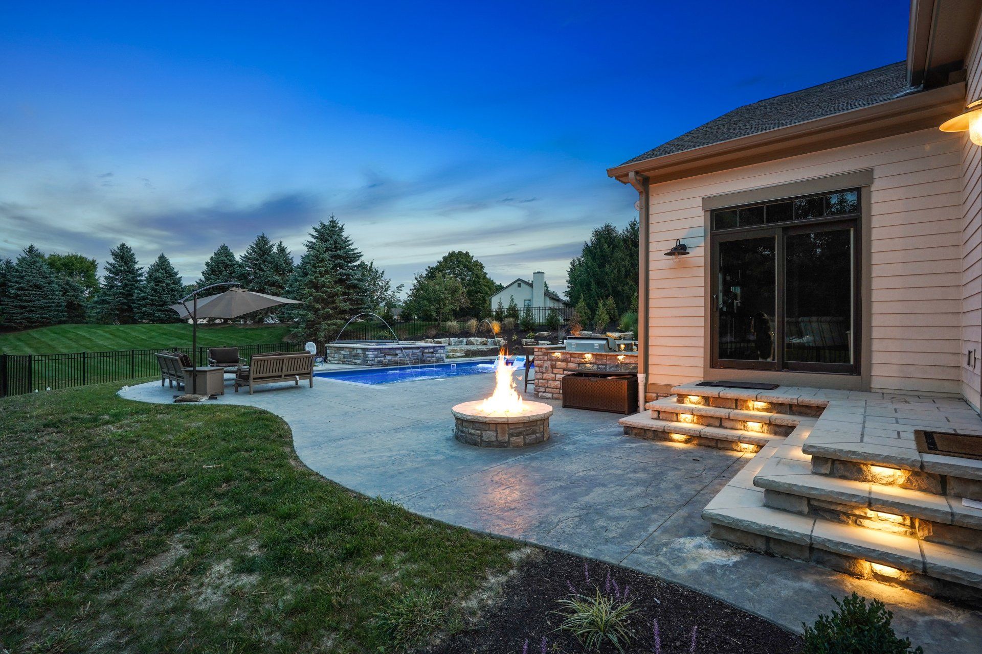 Backyard patio at dusk with fire pit, lit steps, pool, and house exterior