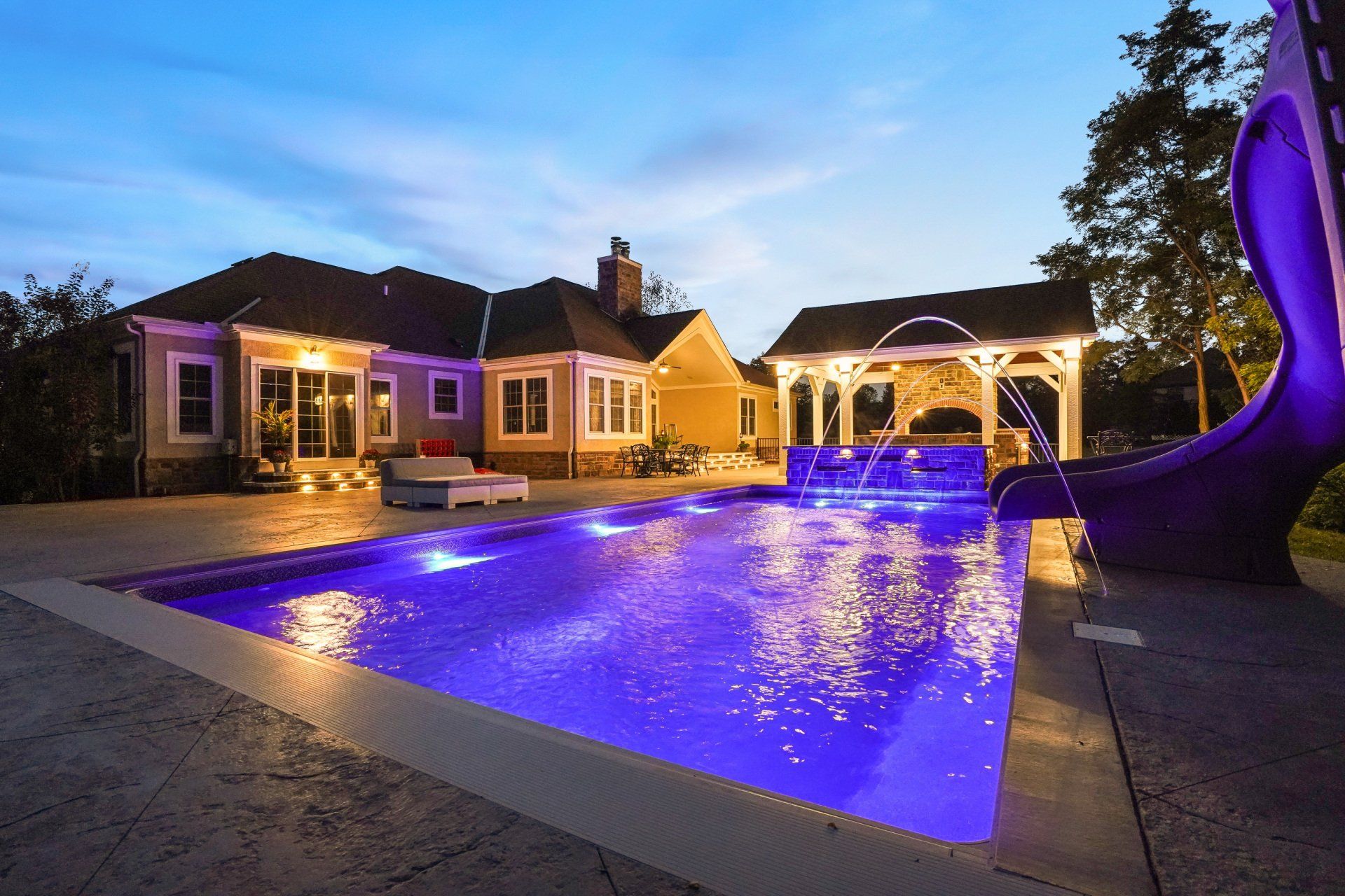 Luxury backyard pool lit blue at dusk beside a house and gazebo