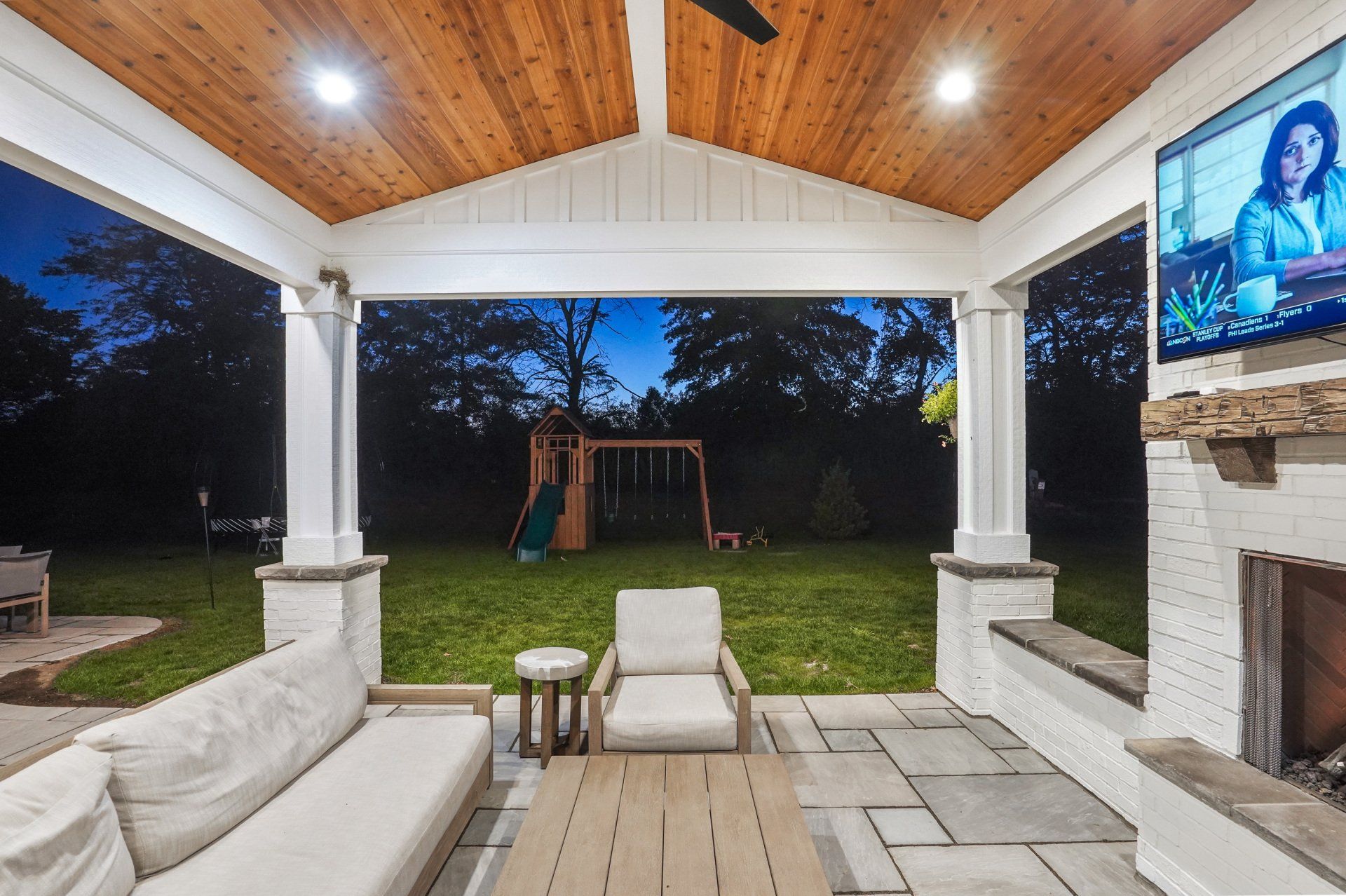Covered patio with white columns, cushioned chairs, TV, and a view of a backyard swing set at dusk