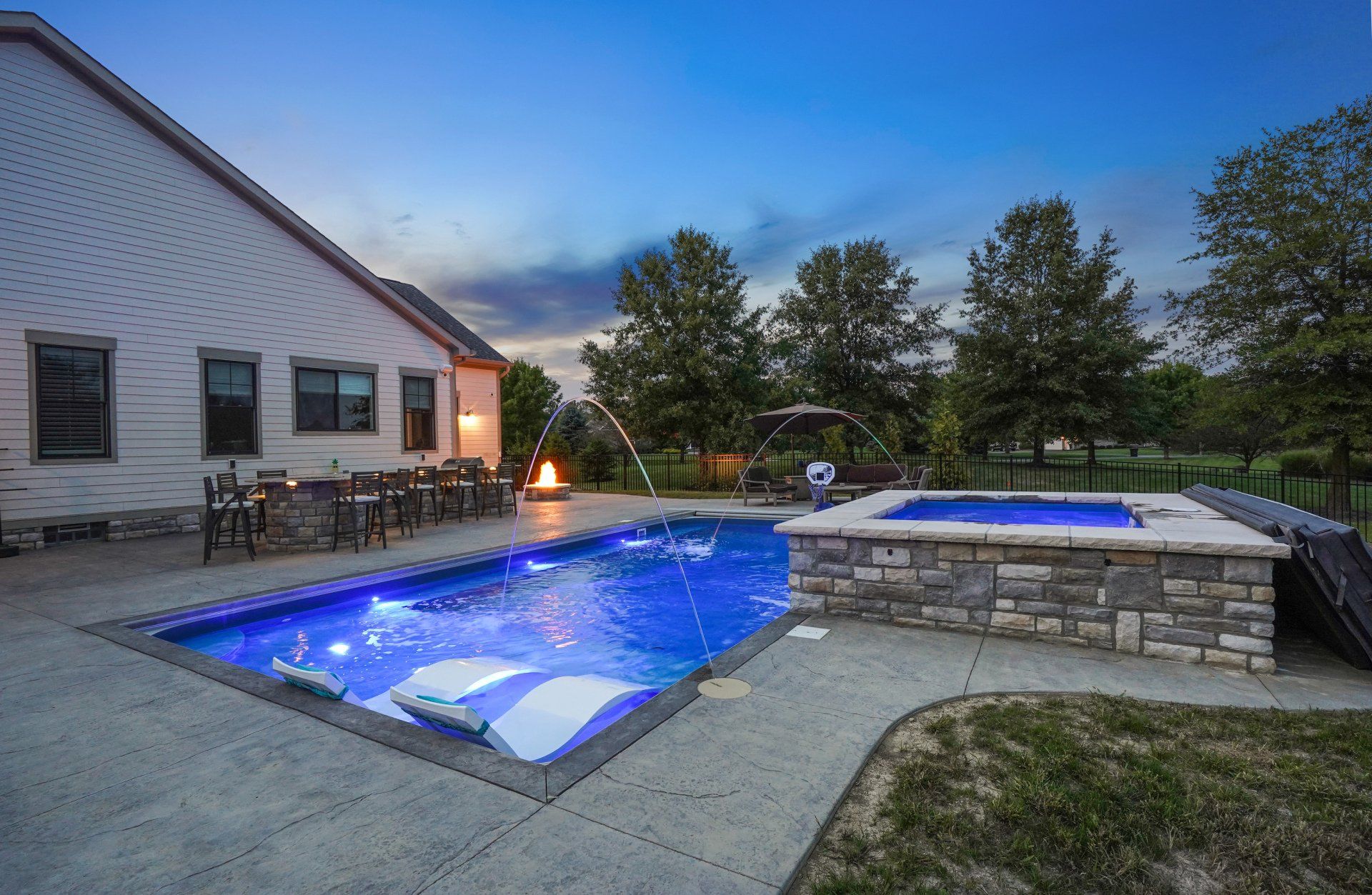 Backyard pool with blue-lit water and a raised hot tub beside a house at dusk