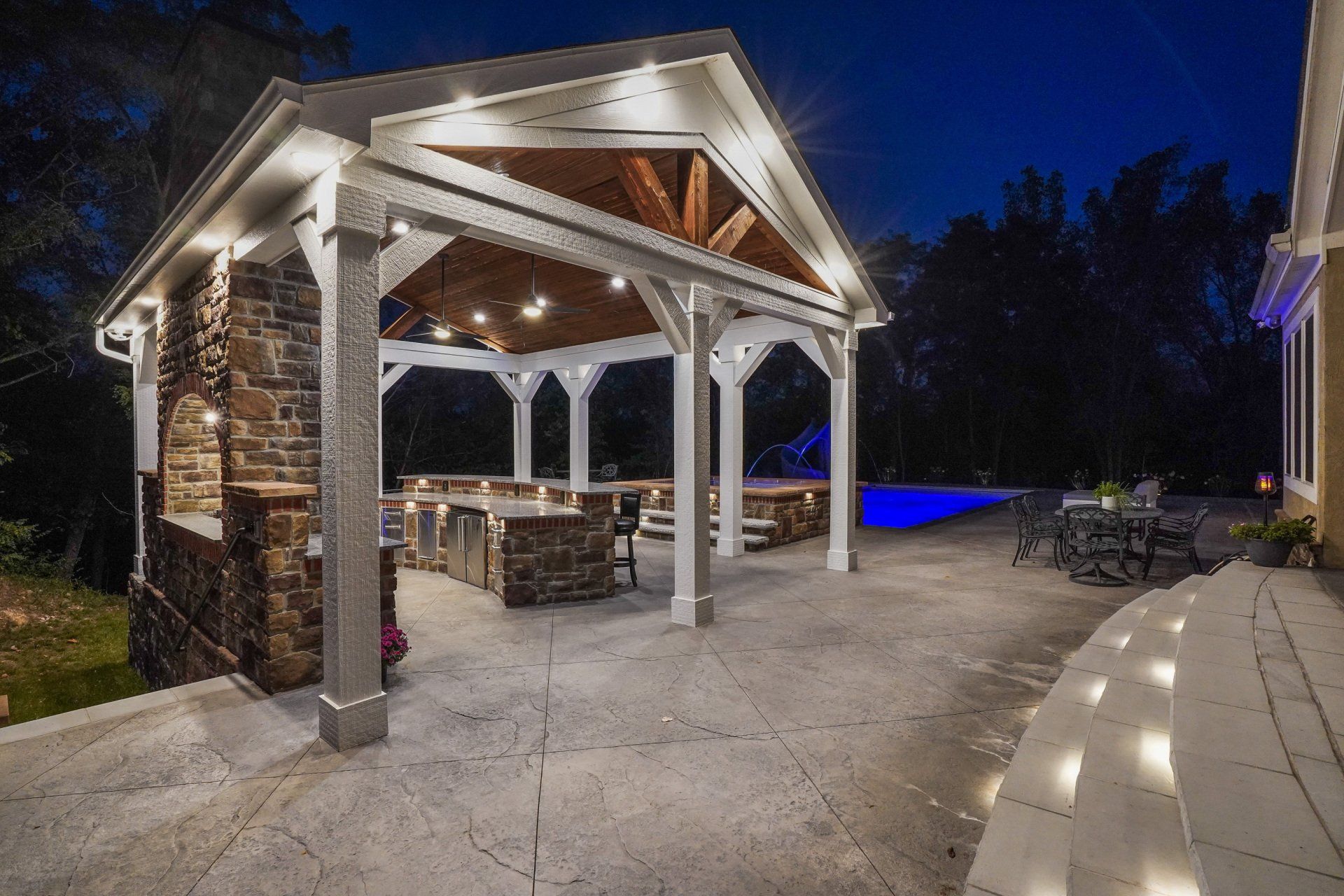 Covered stone patio with white posts and string lights at night, beside a pool and landscaped yard.