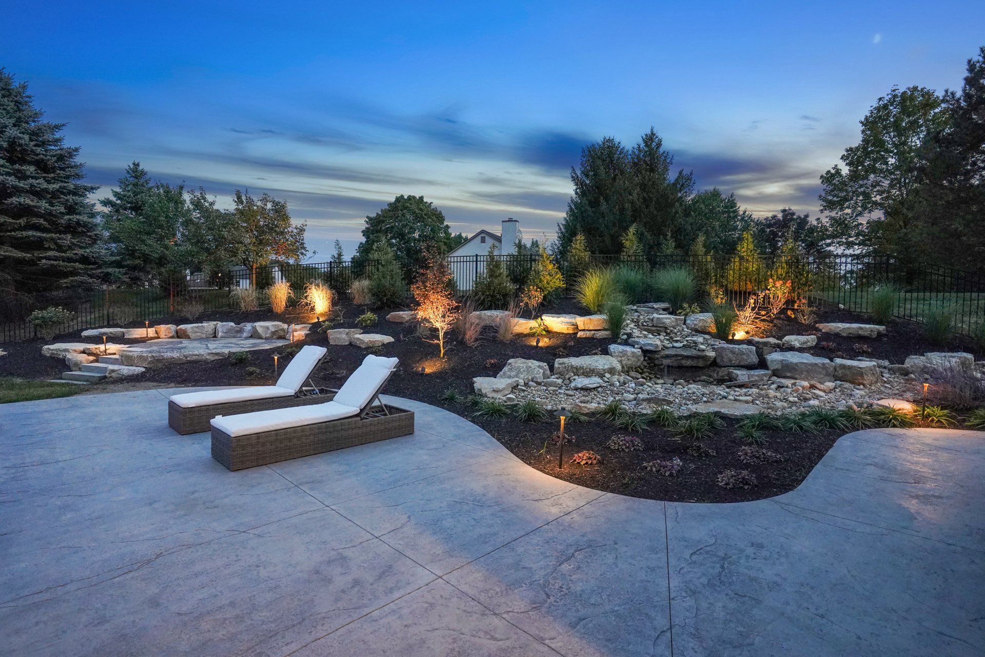 Modern backyard patio with lounge chairs, stone fire pits, and warm landscape lighting at dusk