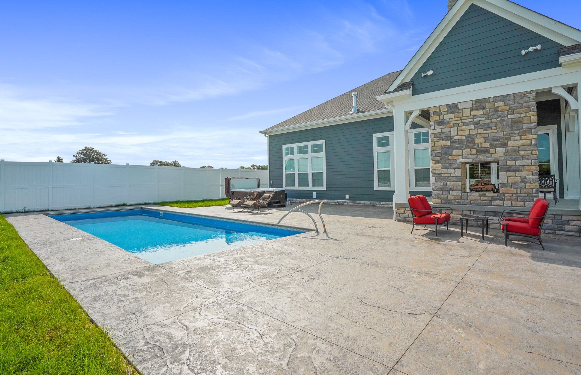 Backyard patio with a swimming pool, concrete deck, stone house, and red patio chairs under a blue sky