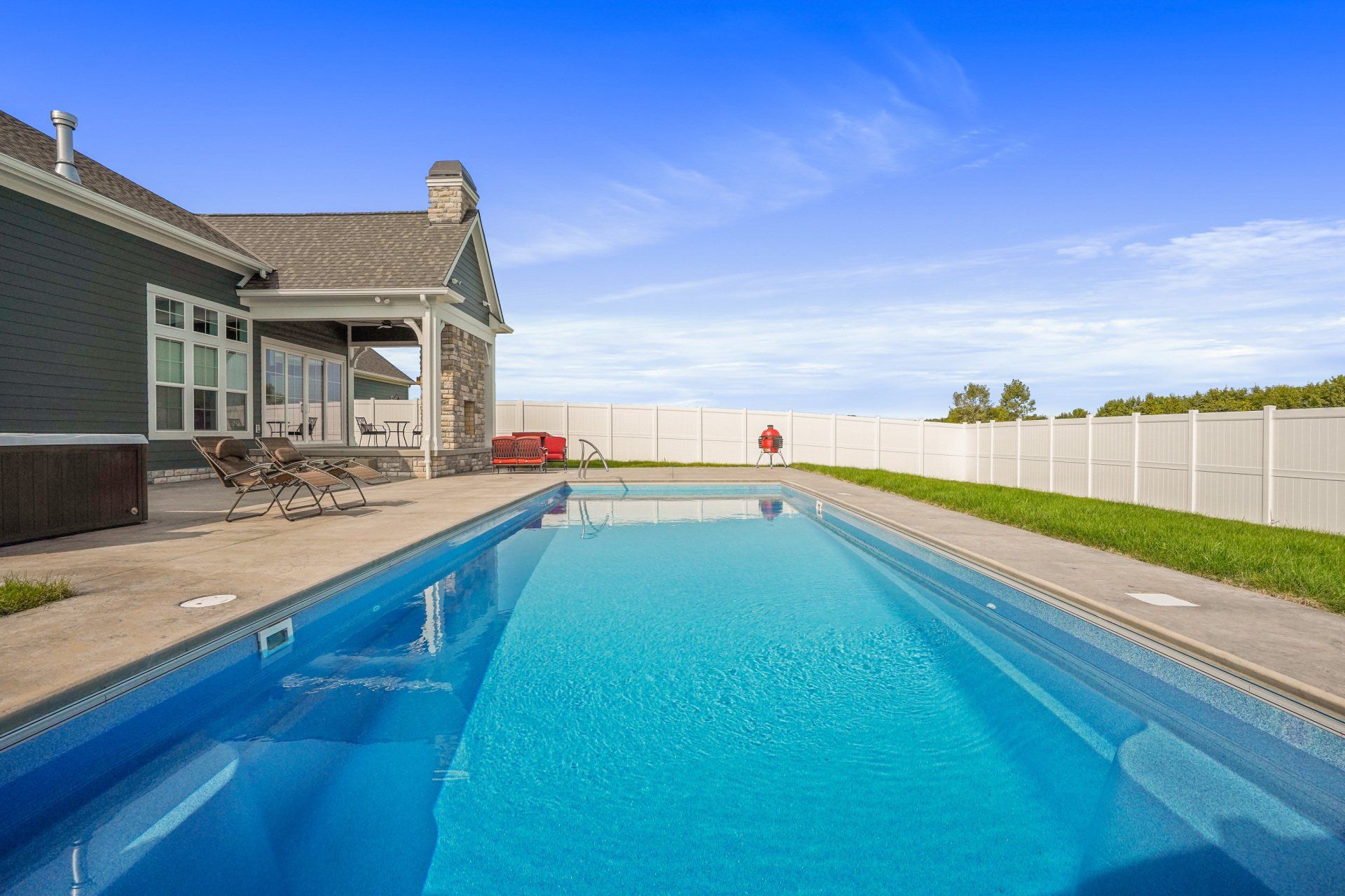 Backyard swimming pool beside a house, with a patio and white fence under a clear blue sky