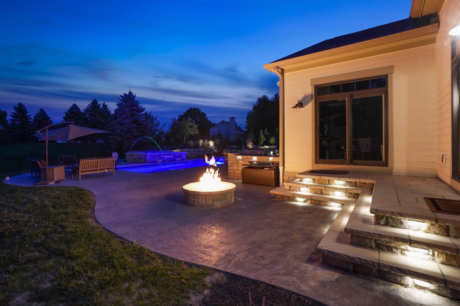 Backyard patio at dusk with lit steps, fire pit, and a house exterior glowing under blue evening sky