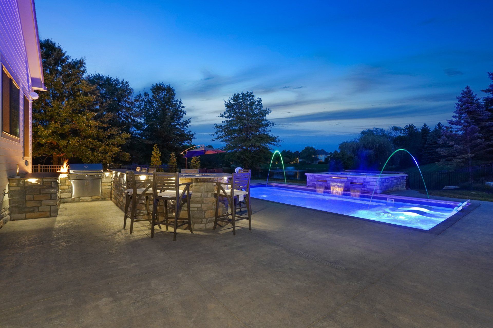 Patio at dusk with outdoor dining set beside a blue-lit swimming pool and waterfall fountain