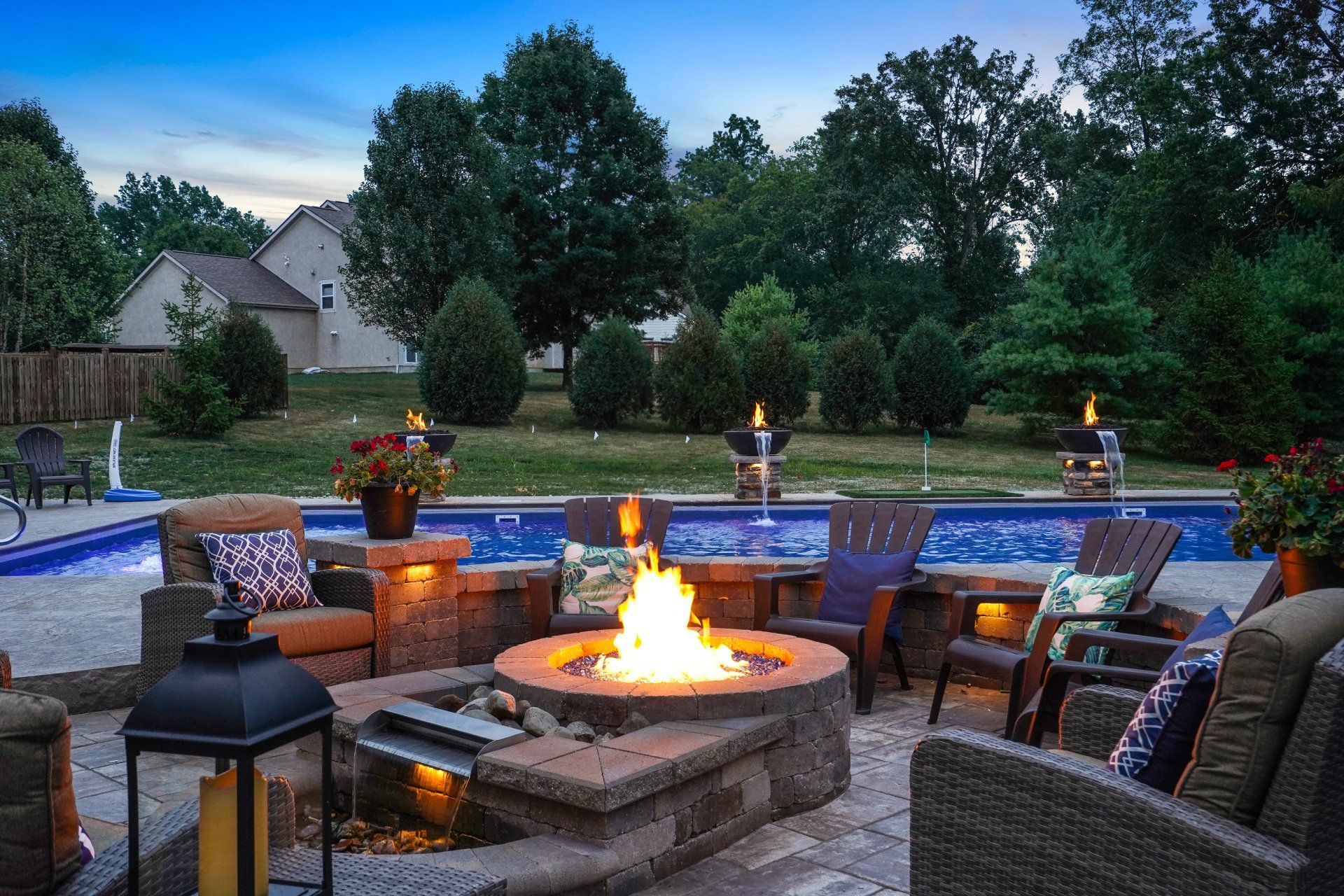 Backyard patio with a stone fire pit, wicker seating, lanterns, and a lit blue-lit pool at dusk