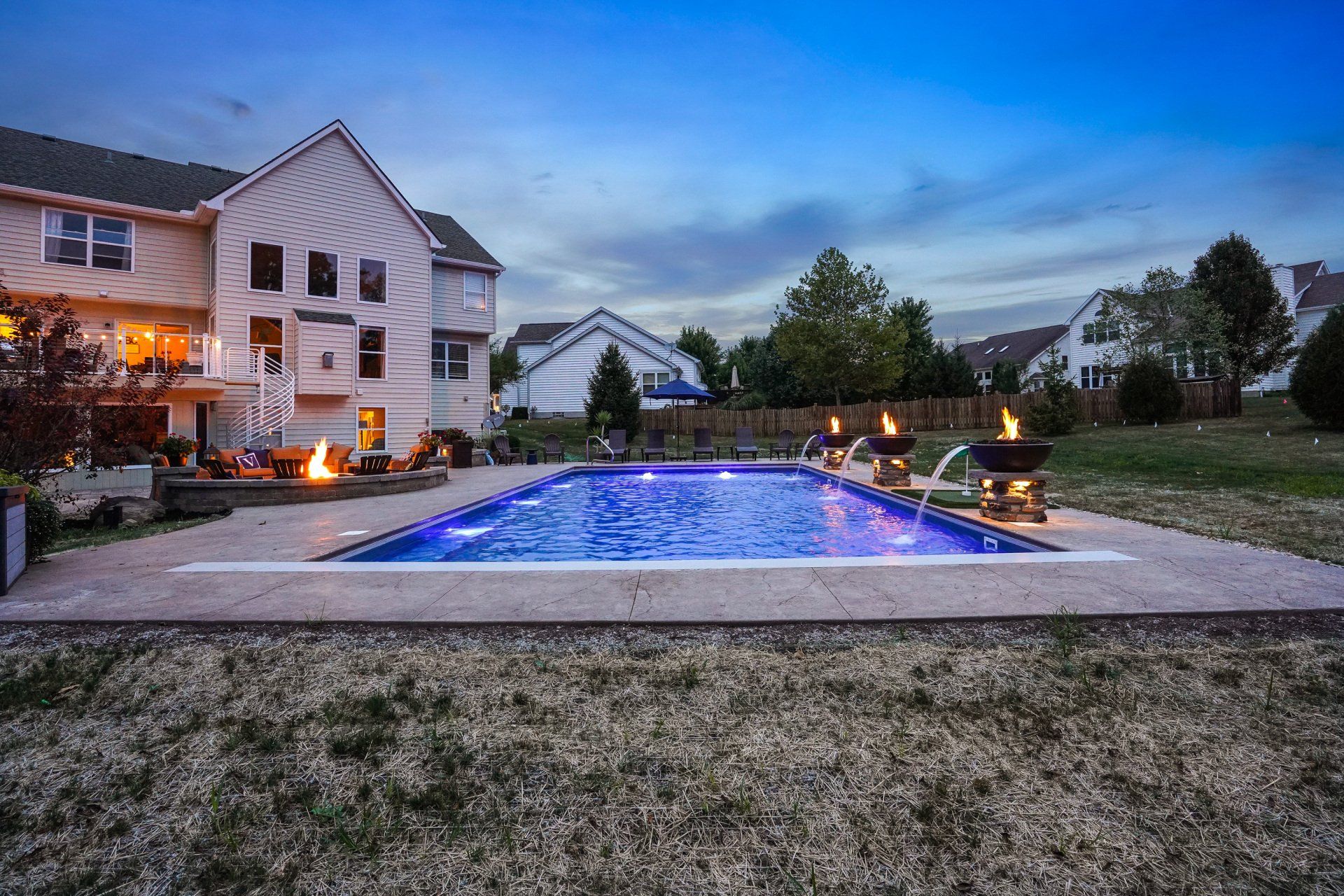 Backyard at dusk with a lit blue swimming pool beside a house and patio seating