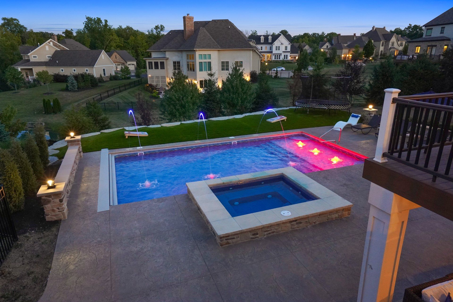 Backyard with lit swimming pool and spa at dusk, surrounded by patio and suburban homes