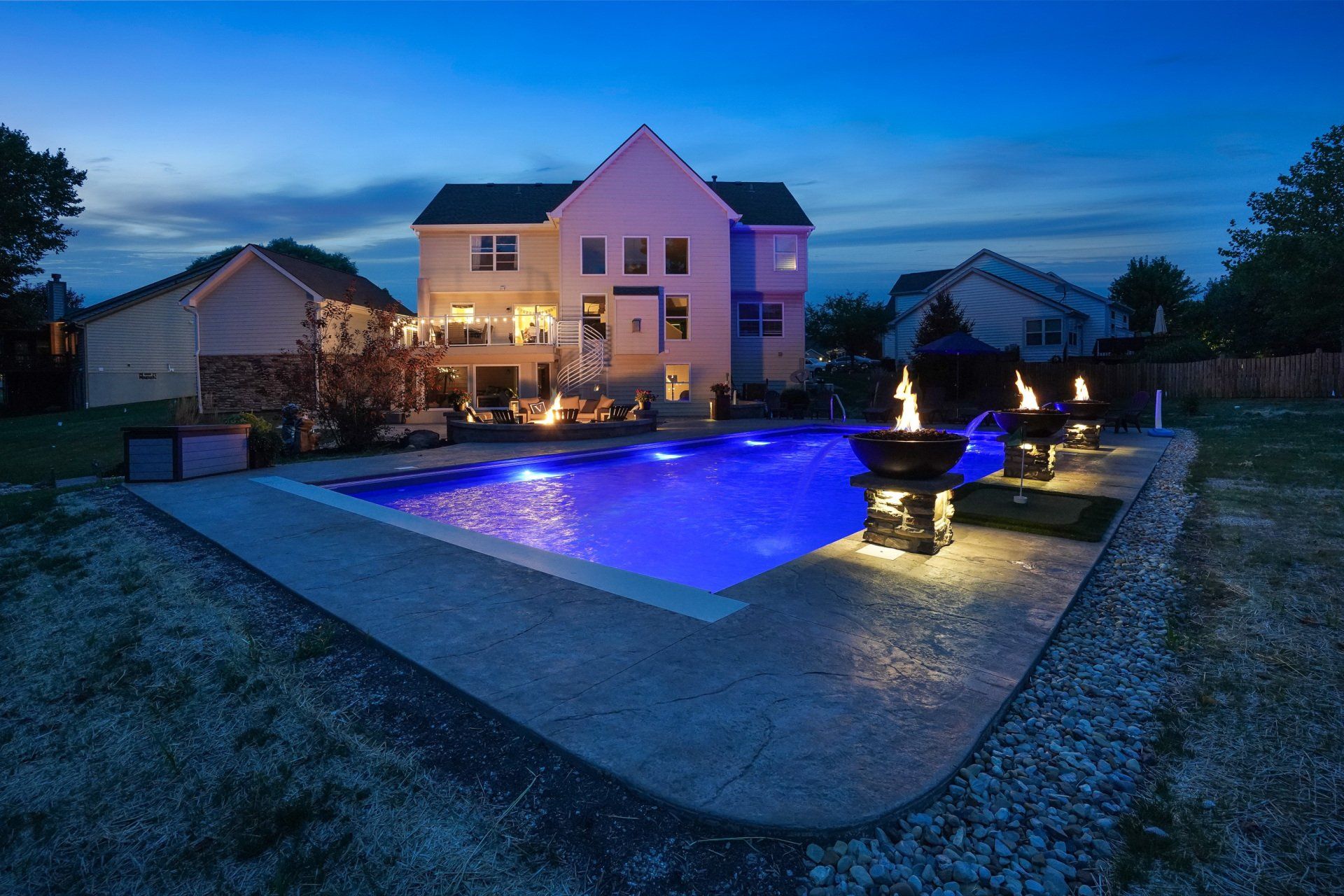 Backyard pool lit blue at dusk beside a large house with warm lights and patio fountains