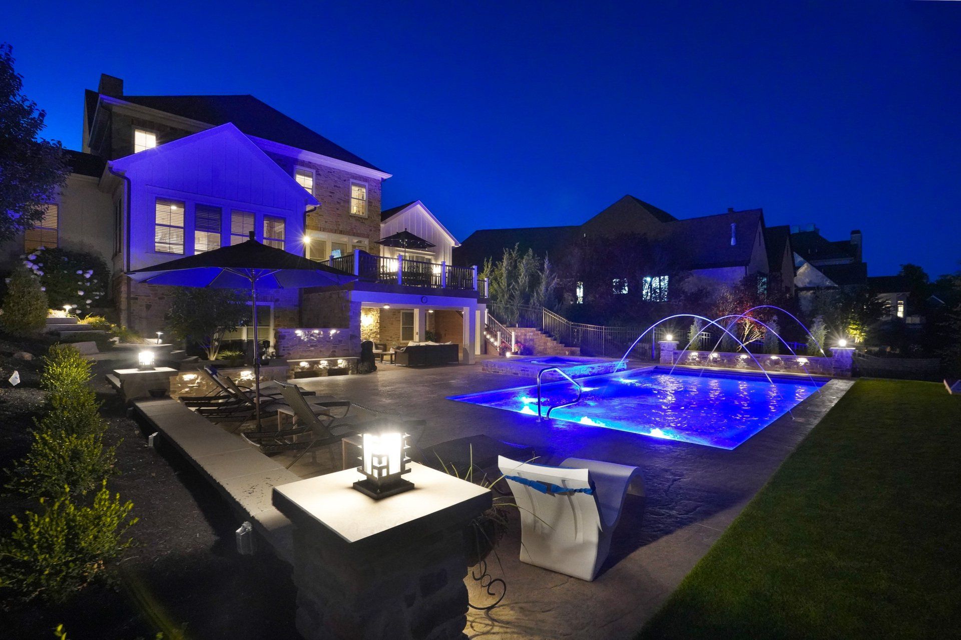 Luxury backyard with a lit blue swimming pool, patio seating, and a modern house at night