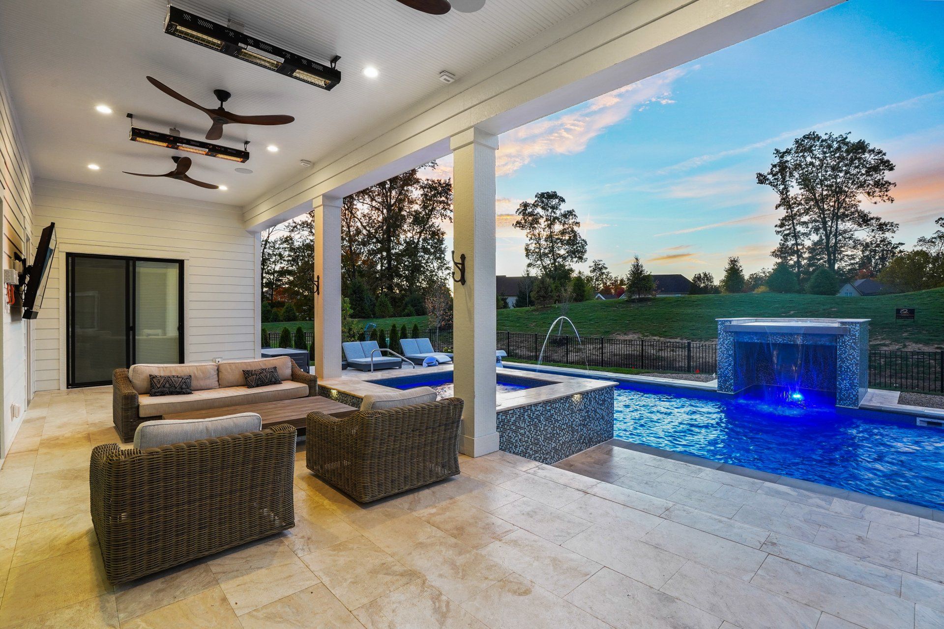 Covered patio with wicker seating overlooking a blue-lit pool and sunset backyard view