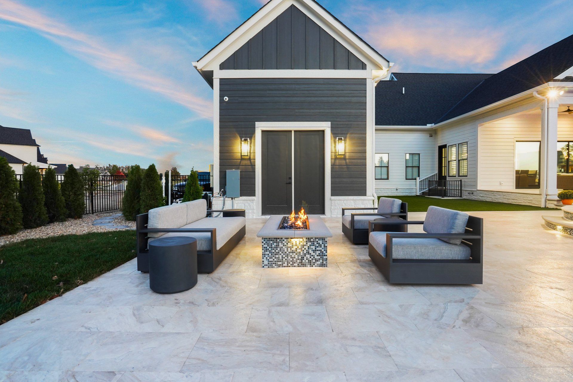 Modern patio with gray outdoor seating, fire pit, and blue-gray house exterior at dusk