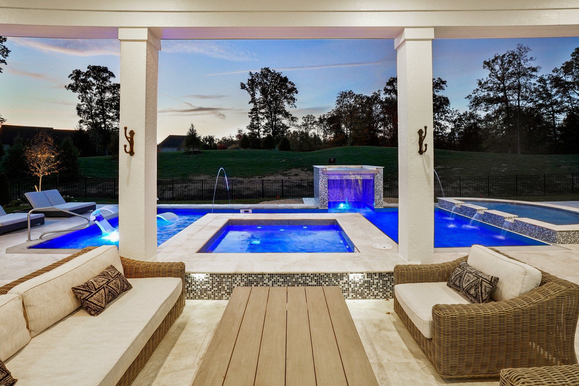 Patio view of a lit blue pool and spa at dusk, framed by a white pergola and wicker seating.