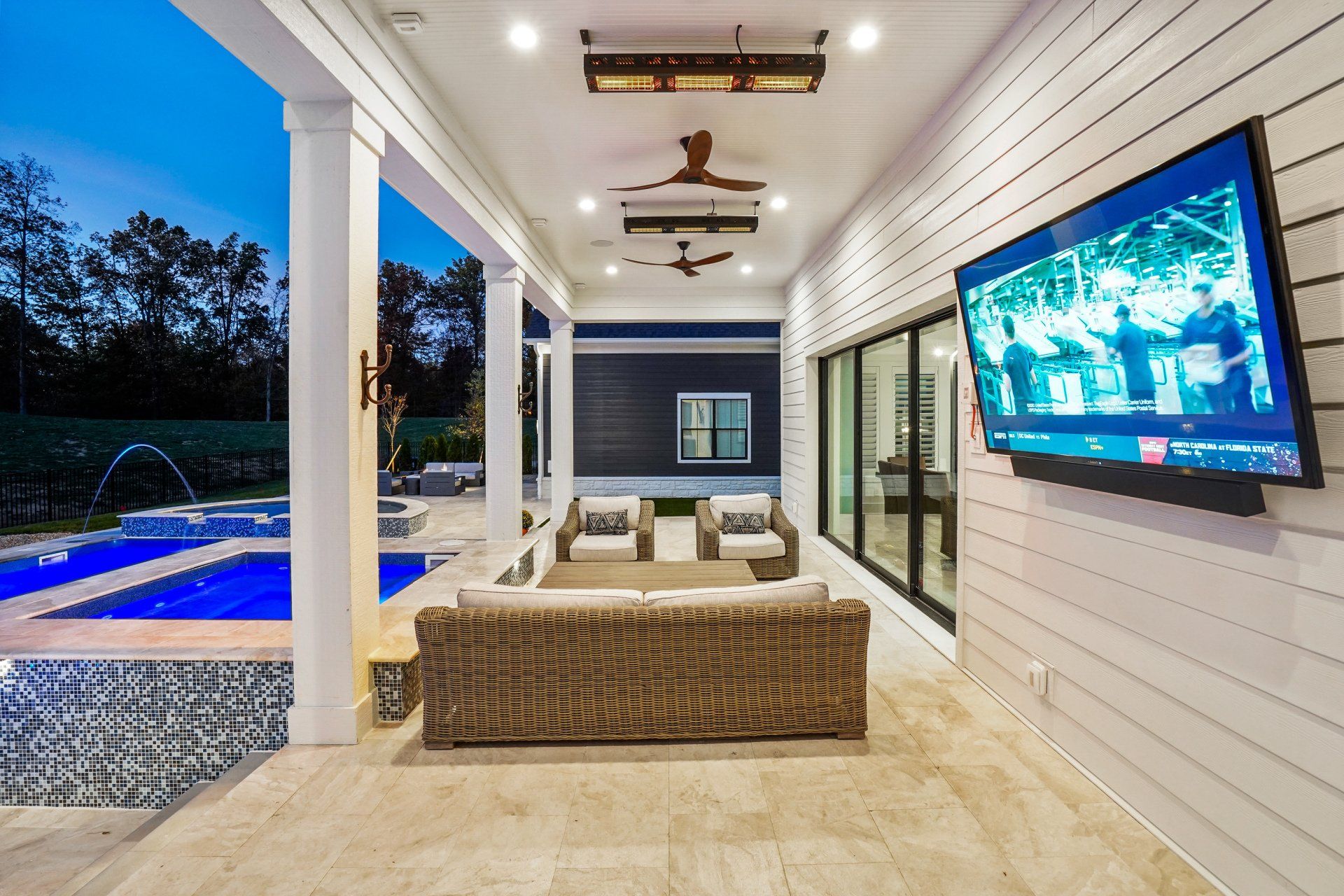 Covered patio with wicker seating, wall-mounted TV, pool view, and ceiling fans at dusk