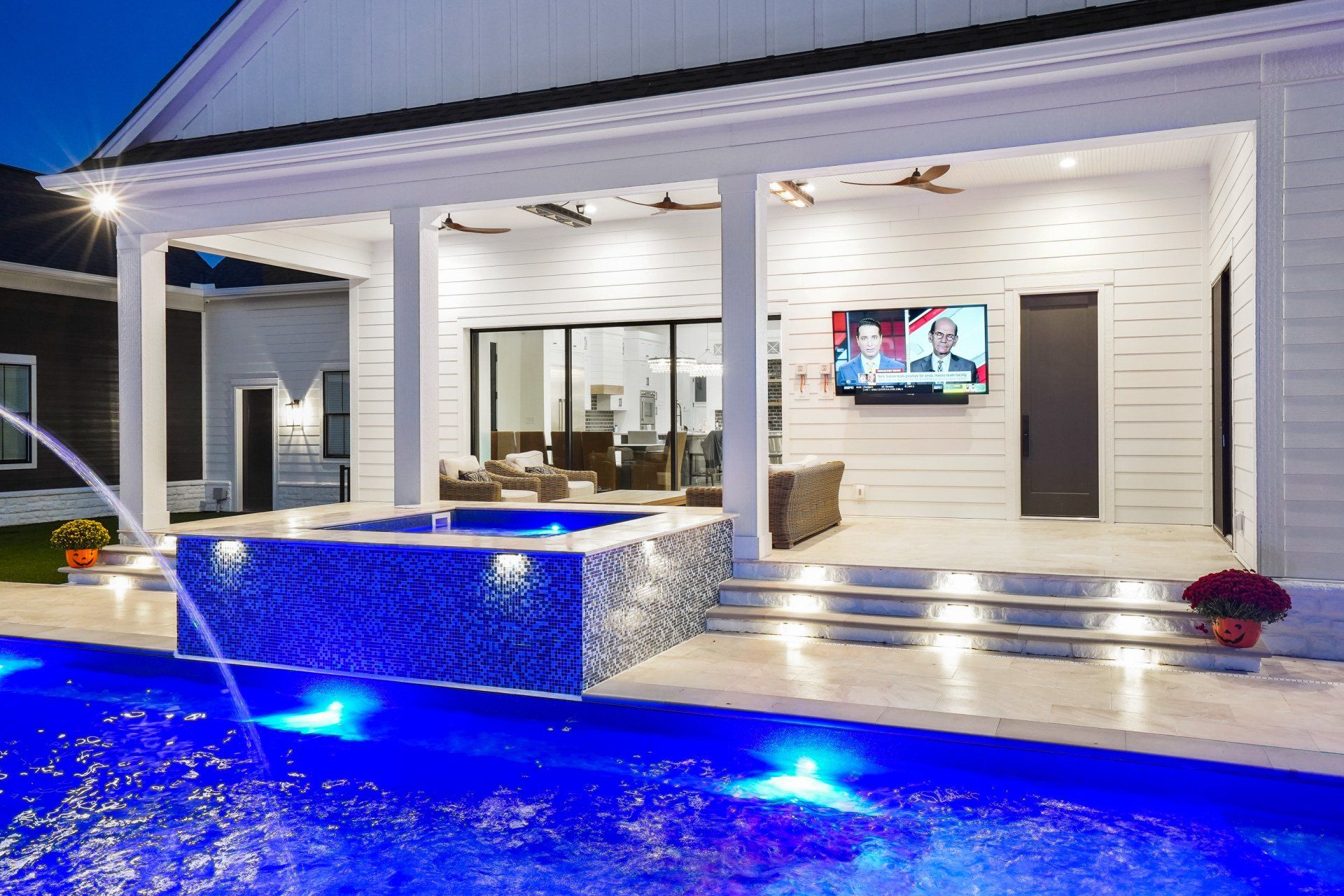 Modern white patio with lit pool, tiled water feature, and a TV visible through large sliding doors at dusk
