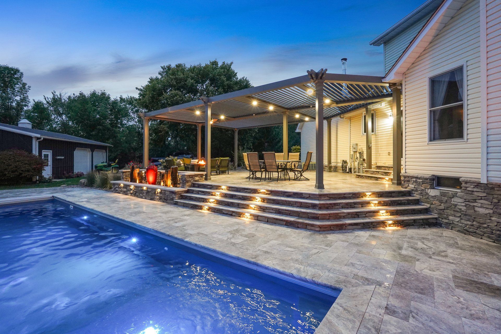 Backyard pool with lit deck, pergola, and house patio at dusk