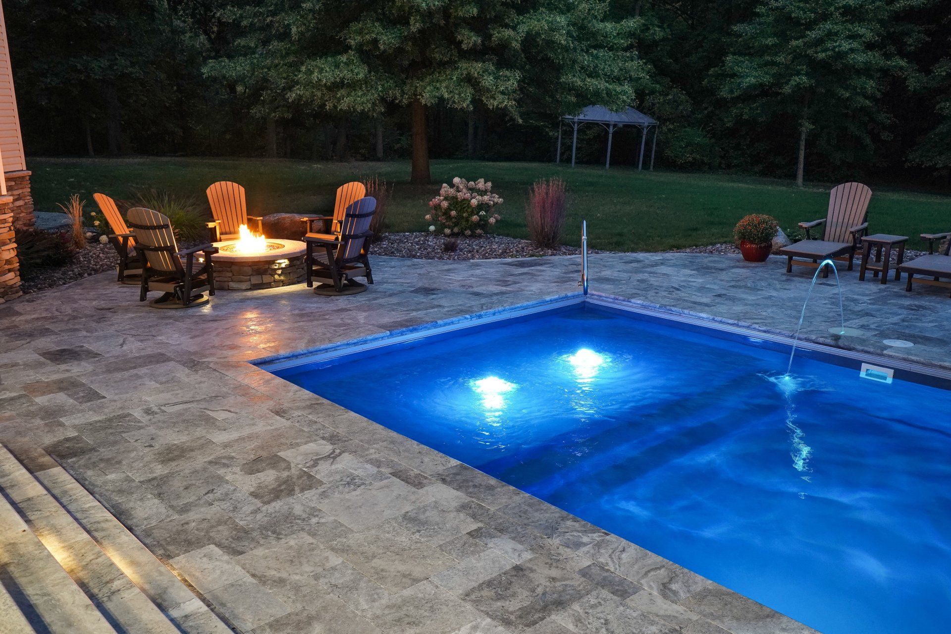 Backyard pool at dusk with glowing blue water and a lit fire pit on a stone patio
