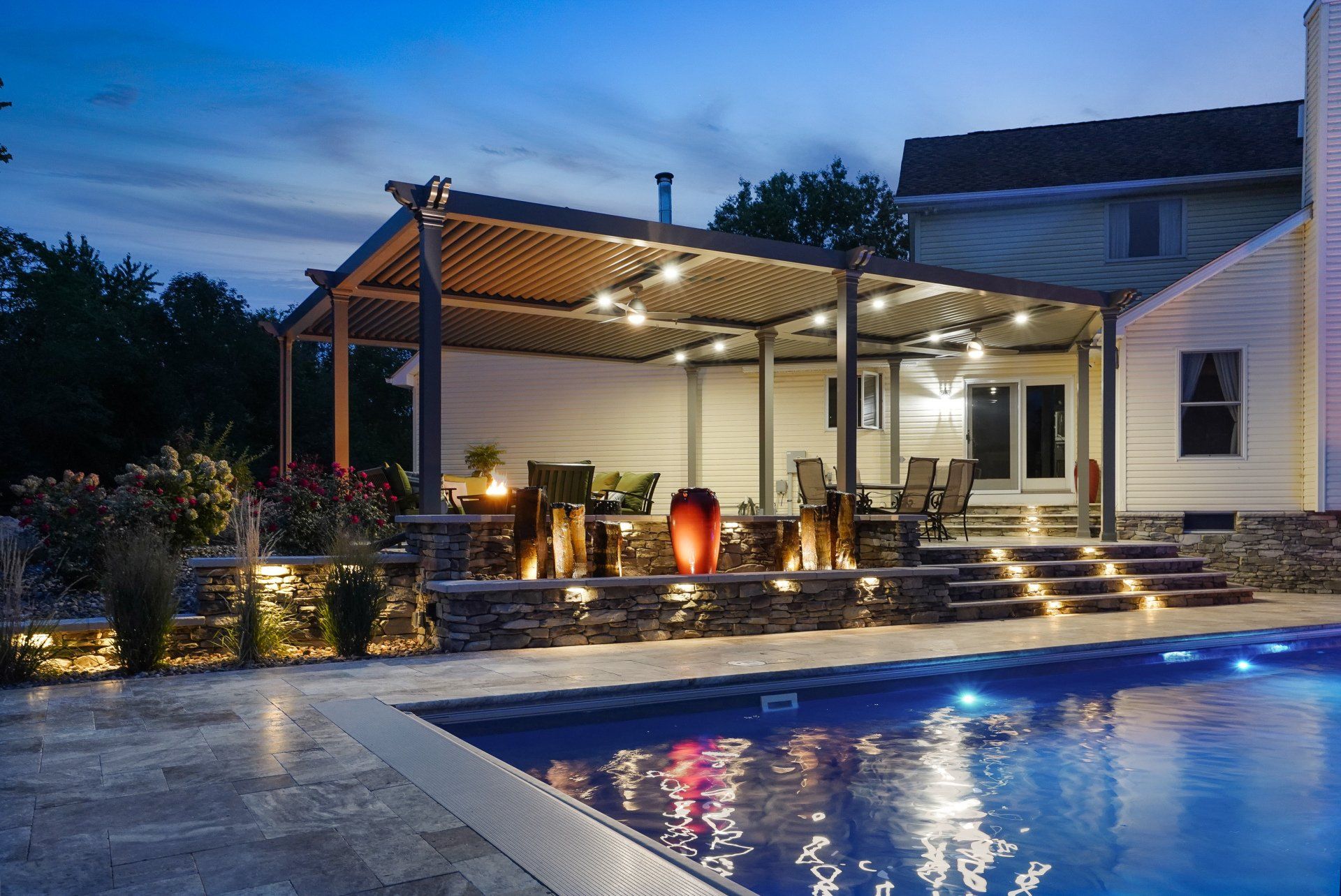 Lit patio beside a glowing pool at dusk, with a covered outdoor dining area and steps into the water